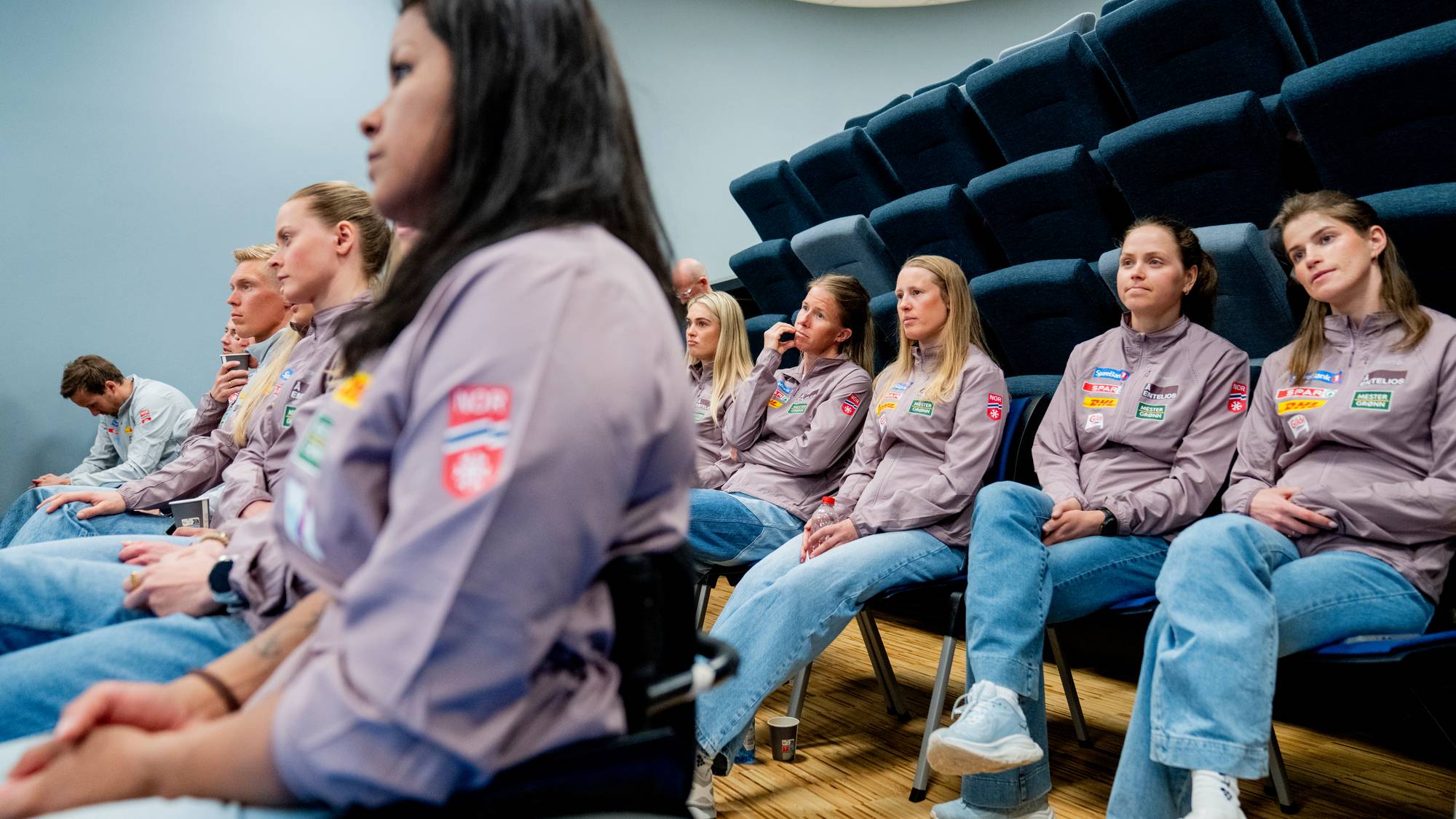 En gruppe personer sitter og ser fremover i en forelesningssal. Mange av dem har på seg like lilla jakker. Bildet er tatt fra siden, med fokus på personen i forgrunnen. I bakgrunnen sitter flere kvinner på rekke. (Bildebeskrivelsen er laget av en KI-tjeneste)