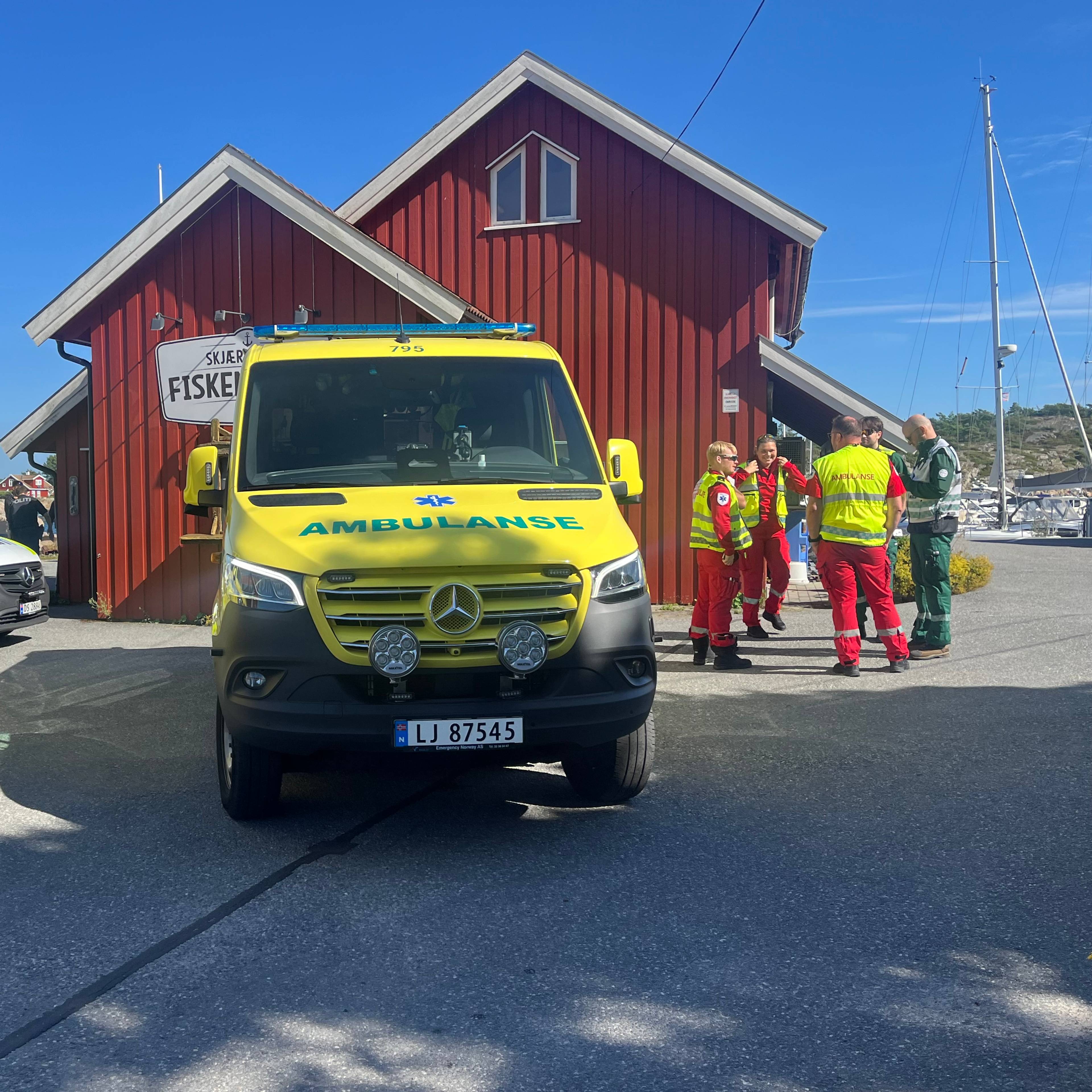 Ambulanser og helsepersonell ved en rød trebygning i en havn. (Bildebeskrivelsen er laget av en KI-tjeneste)