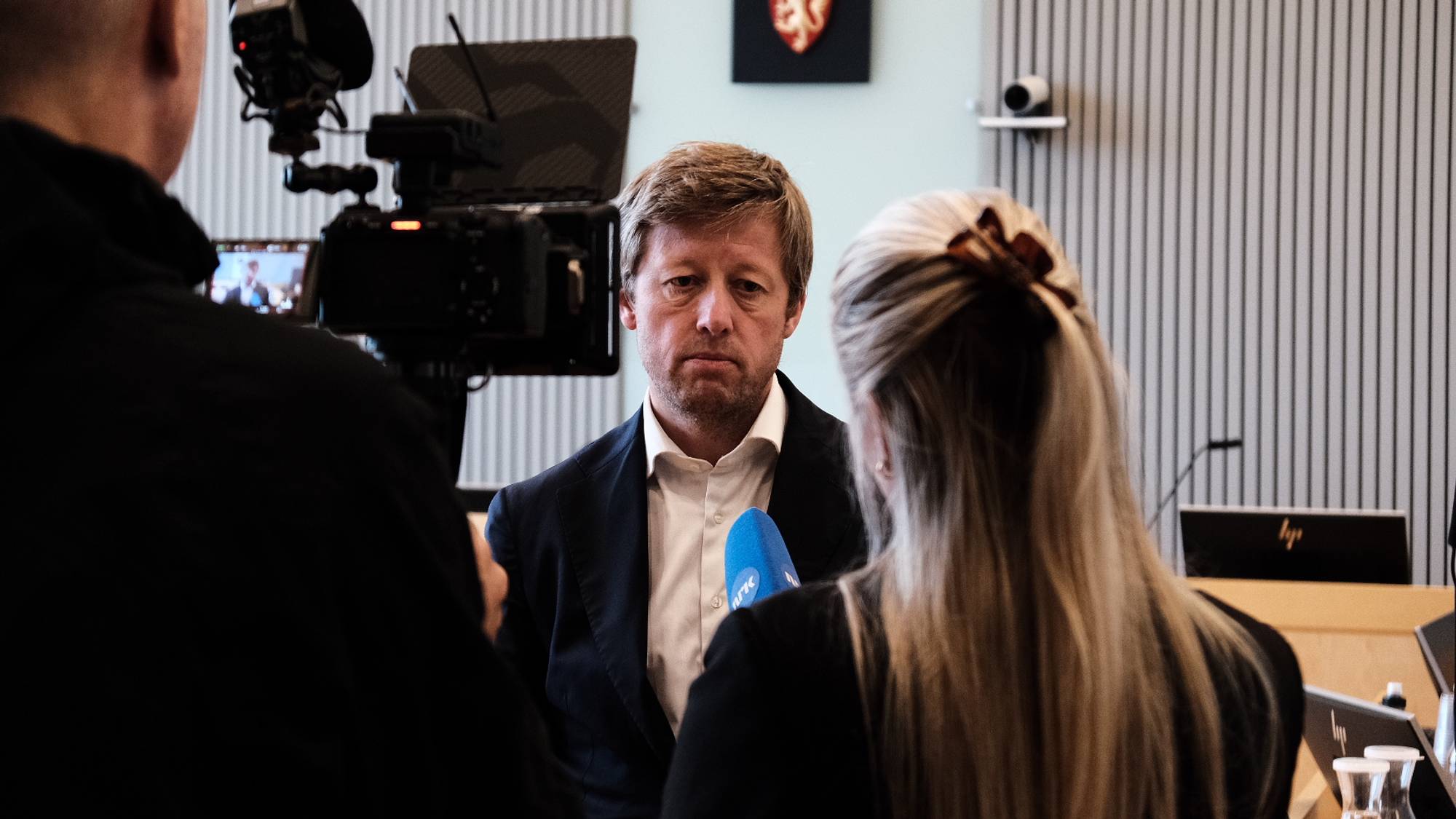 Kvinne intervjuer en mann i dress foran et kamera i en rettssal. Rikssegl på veggen bak. (Bildebeskrivelsen er laget av en KI-tjeneste)
