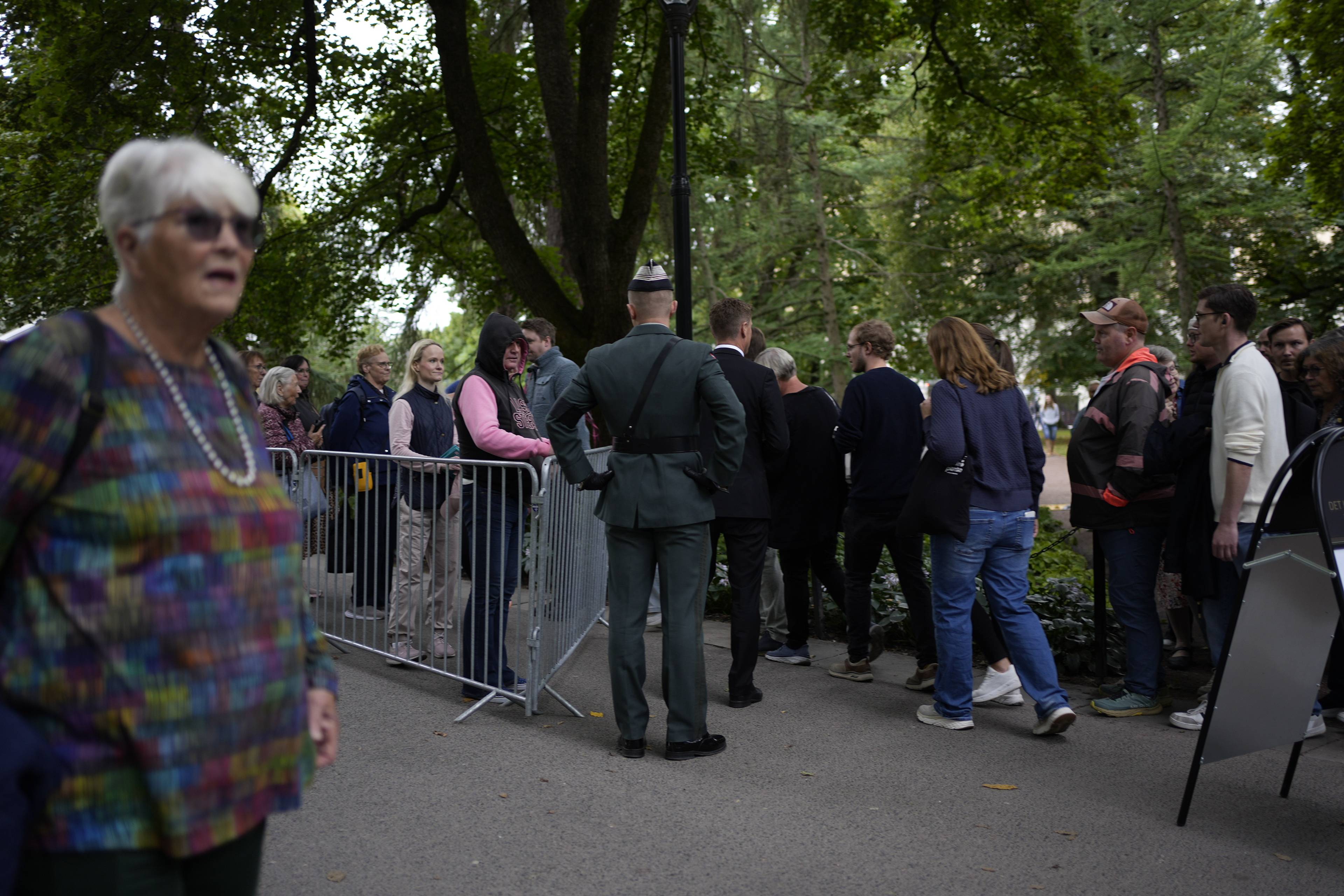 Folk står i kø bak et metallgjerde i en park med en uniformert vakt i forgrunnen. (Bildebeskrivelsen er laget av en KI-tjeneste)