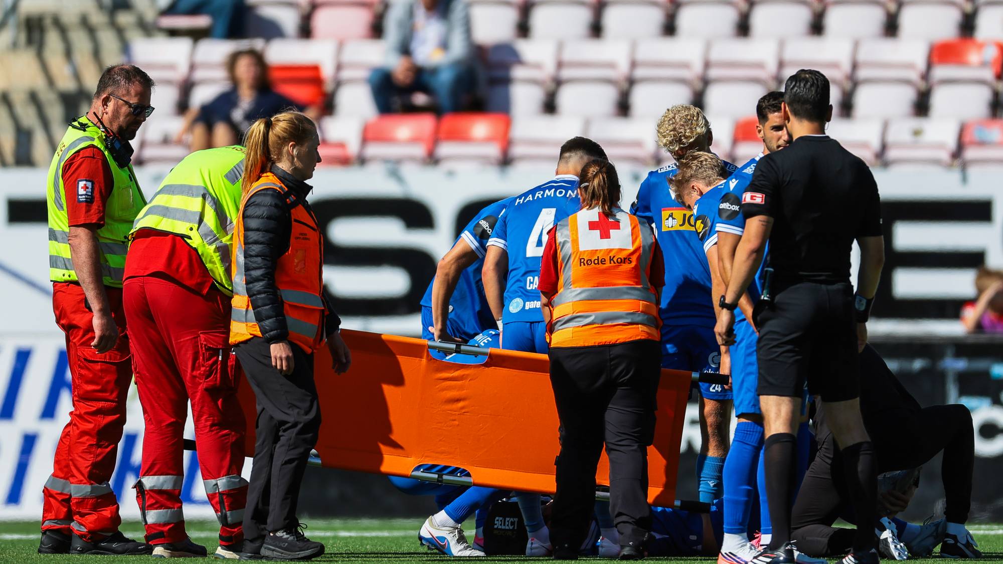 Røde Kors-personell og fotballspillere samlet rundt en skadet spiller på en båre under en kamp. (Bildebeskrivelsen er laget av en KI-tjeneste)