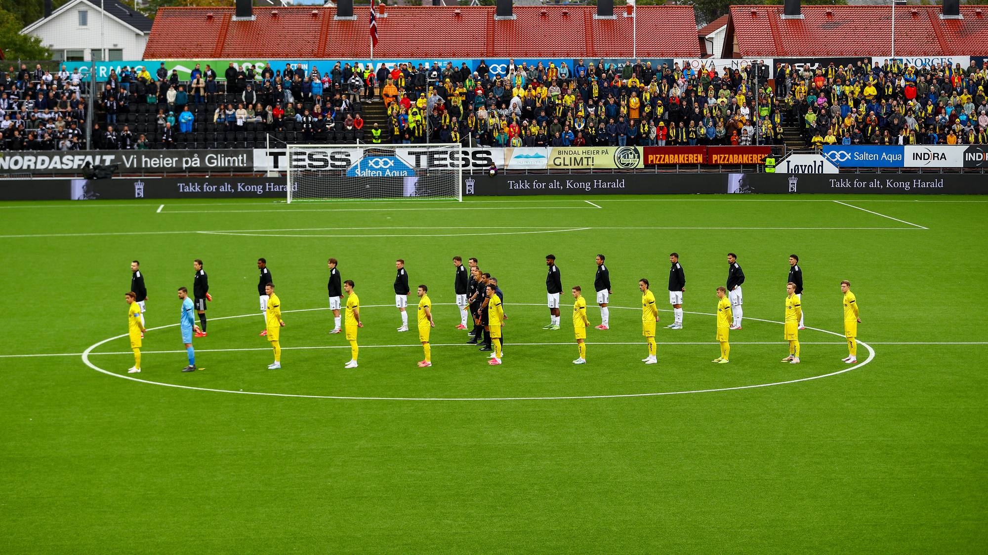 Fotballspillere står oppstilt i en sirkel på banen før kampstart foran et fullsatt publikum. (Bildebeskrivelsen er laget av en KI-tjeneste)