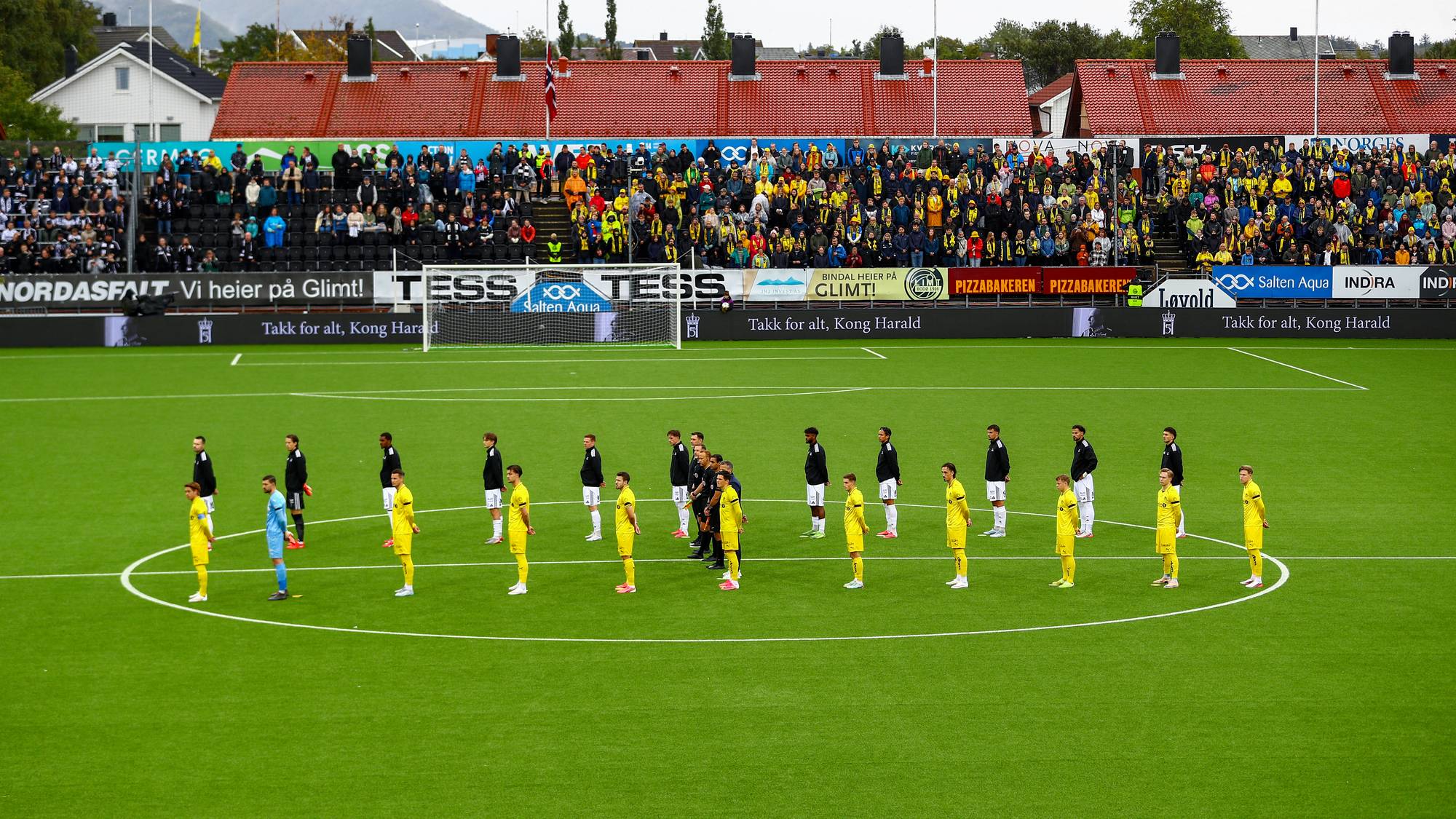 Fotballspillere står oppstilt i en sirkel på banen før kampstart foran et fullsatt publikum. (Bildebeskrivelsen er laget av en KI-tjeneste)