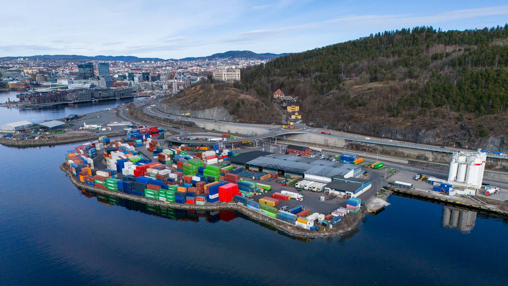 Flyfoto av en stor containerhavn. Mange fargerike containere er stablet på en kai. I bakgrunnen er et bylandskap med bygninger og en motorvei. En skogkledd ås dominerer horisonten. (Bildebeskrivelsen er laget av en KI-tjeneste)
