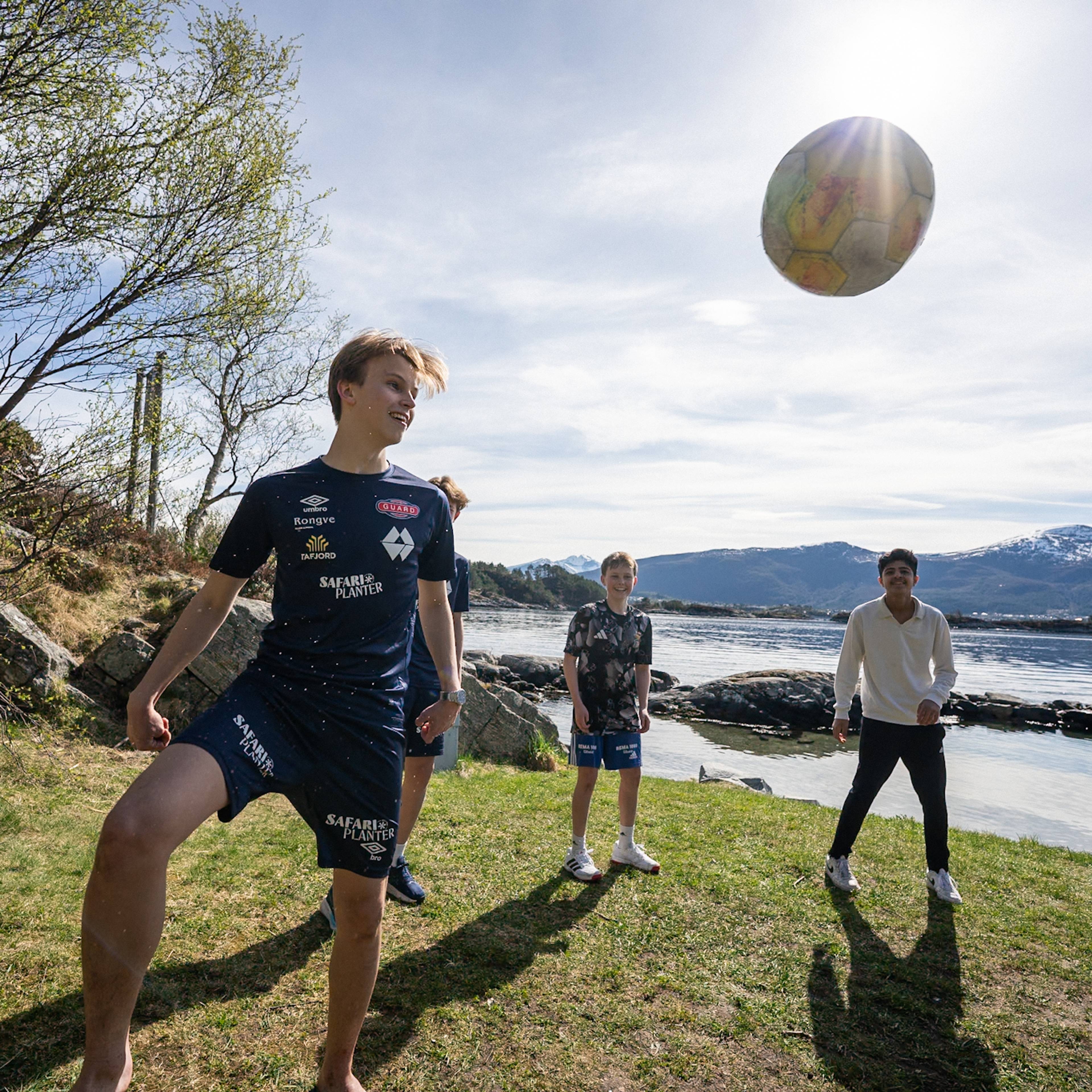 En gruppe gutter spiller fotball utendørs en solrik dag. En av guttene står i forgrunnen og sparker til en fotball som er i luften. Bak ham står flere gutter og ser på. De befinner seg på en gressbakke ved vannet, med fjell i bakgrunnen. (Bildebeskrivelsen er laget av en KI-tjeneste)