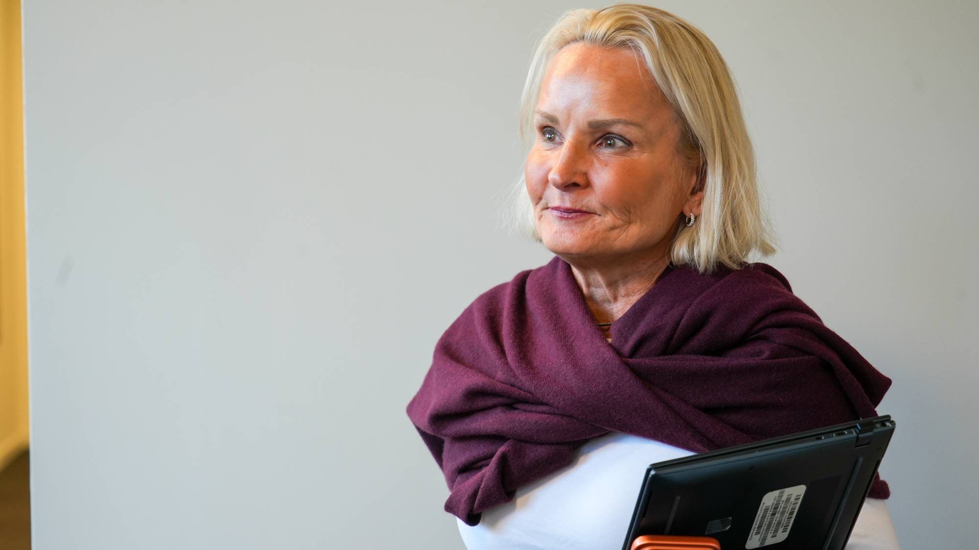 Kvinne med lyst hår og burgunder skjerf holder et nettbrett foran en lys vegg. (Bildebeskrivelsen er laget av en KI-tjeneste)
