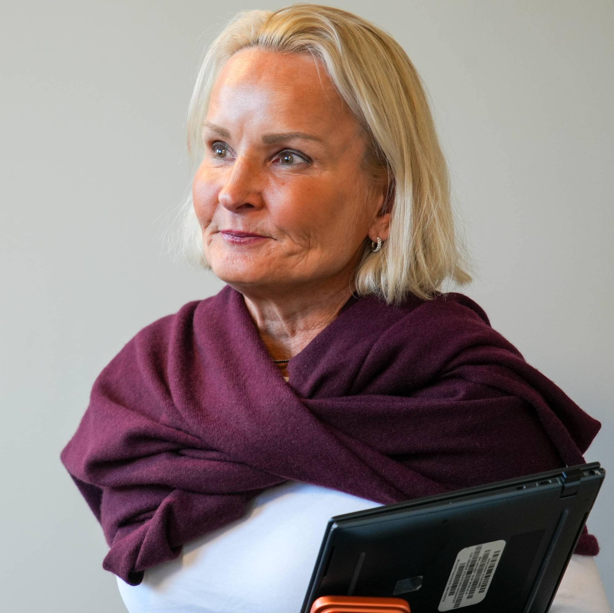 Kvinne med lyst hår og burgunder skjerf holder et nettbrett foran en lys vegg. (Bildebeskrivelsen er laget av en KI-tjeneste)