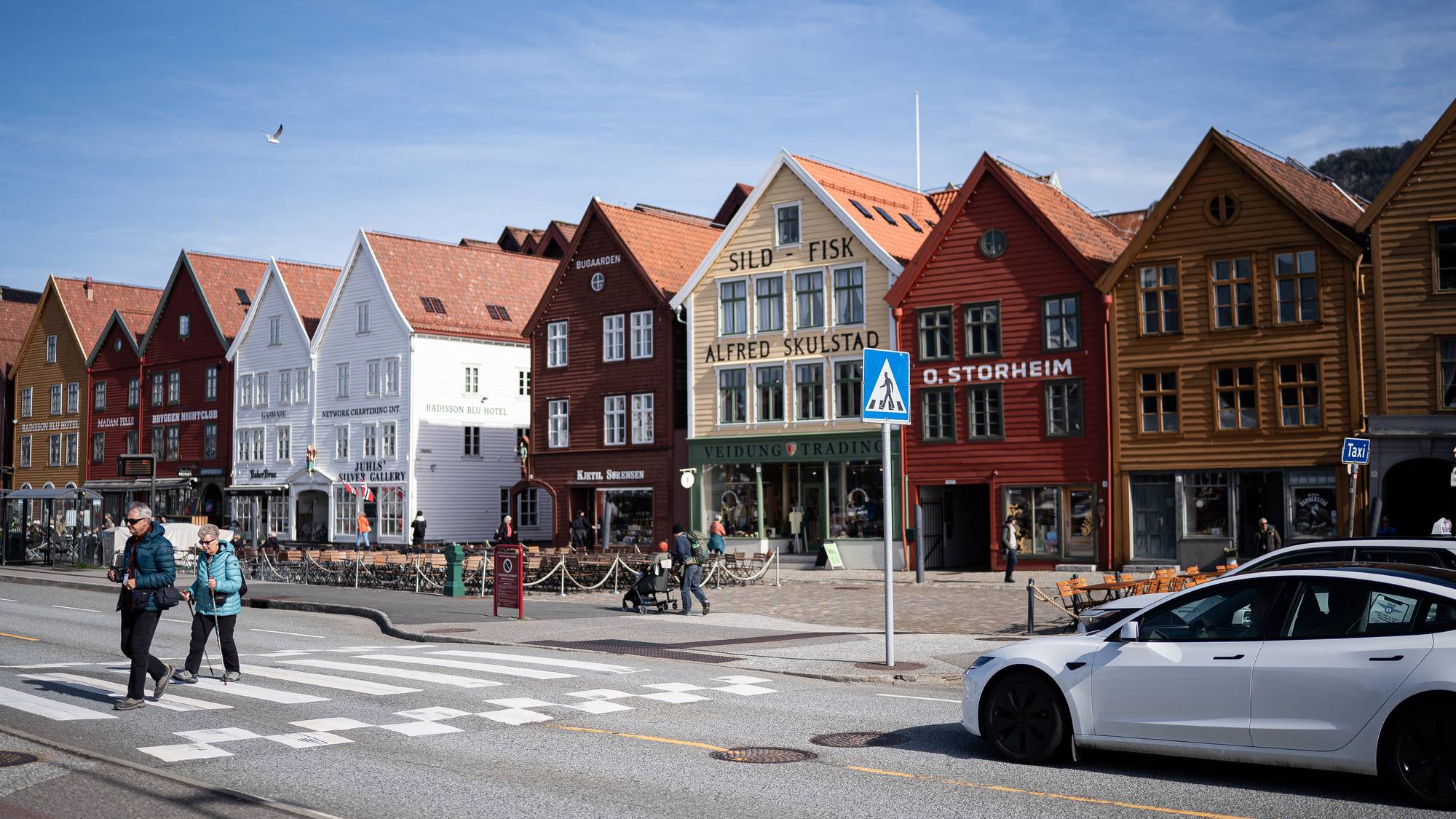 Trehusbebyggelsen på Bryggen i Bergen i sol april 2026. Biler, turister, trafikanter og handlende i vårværet. 