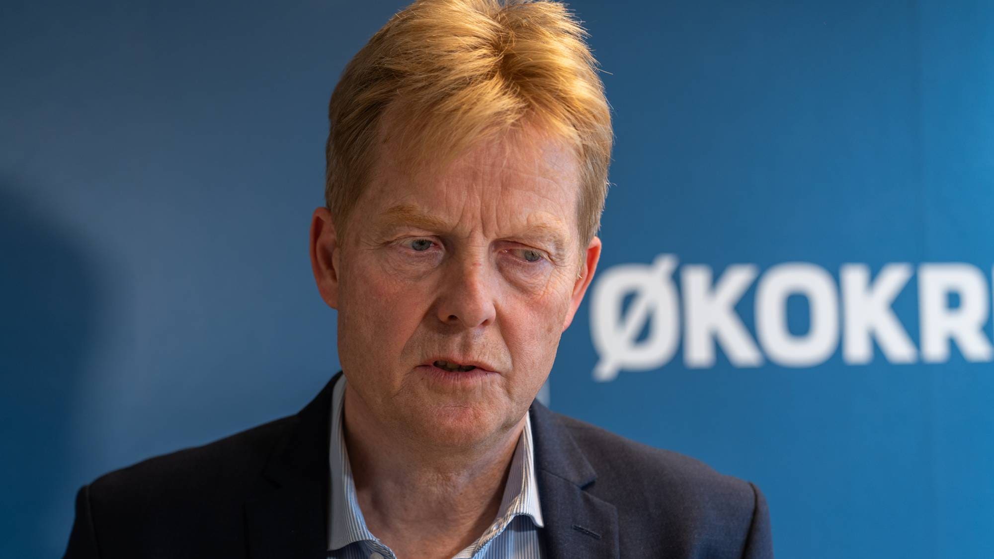 En mann med blondt hår og dress snakker foran en blå bakgrunn der det står Økokrim