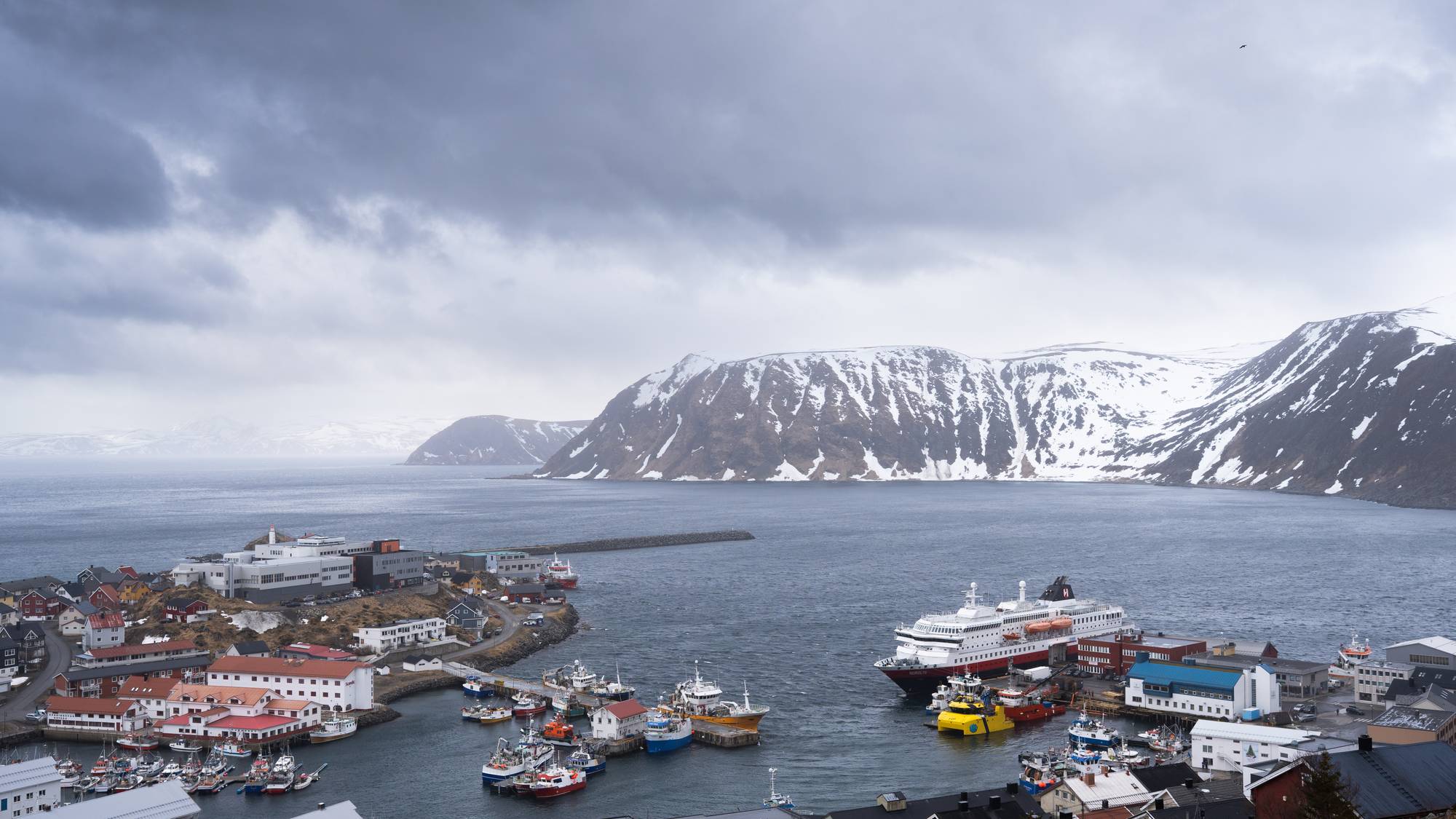 Oversiktsbilde av Honningsvåg sentrum tatt fra fjellet. Båter i havna og hurtigruta MS Nordlys ved kai. Grå himmel og snø i fjella