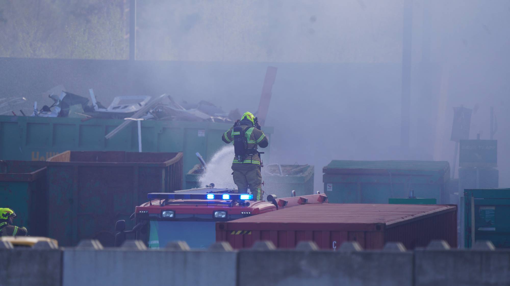 En brannmann står på taket til en brannbil og holder en brannslange han spyler på containere.
