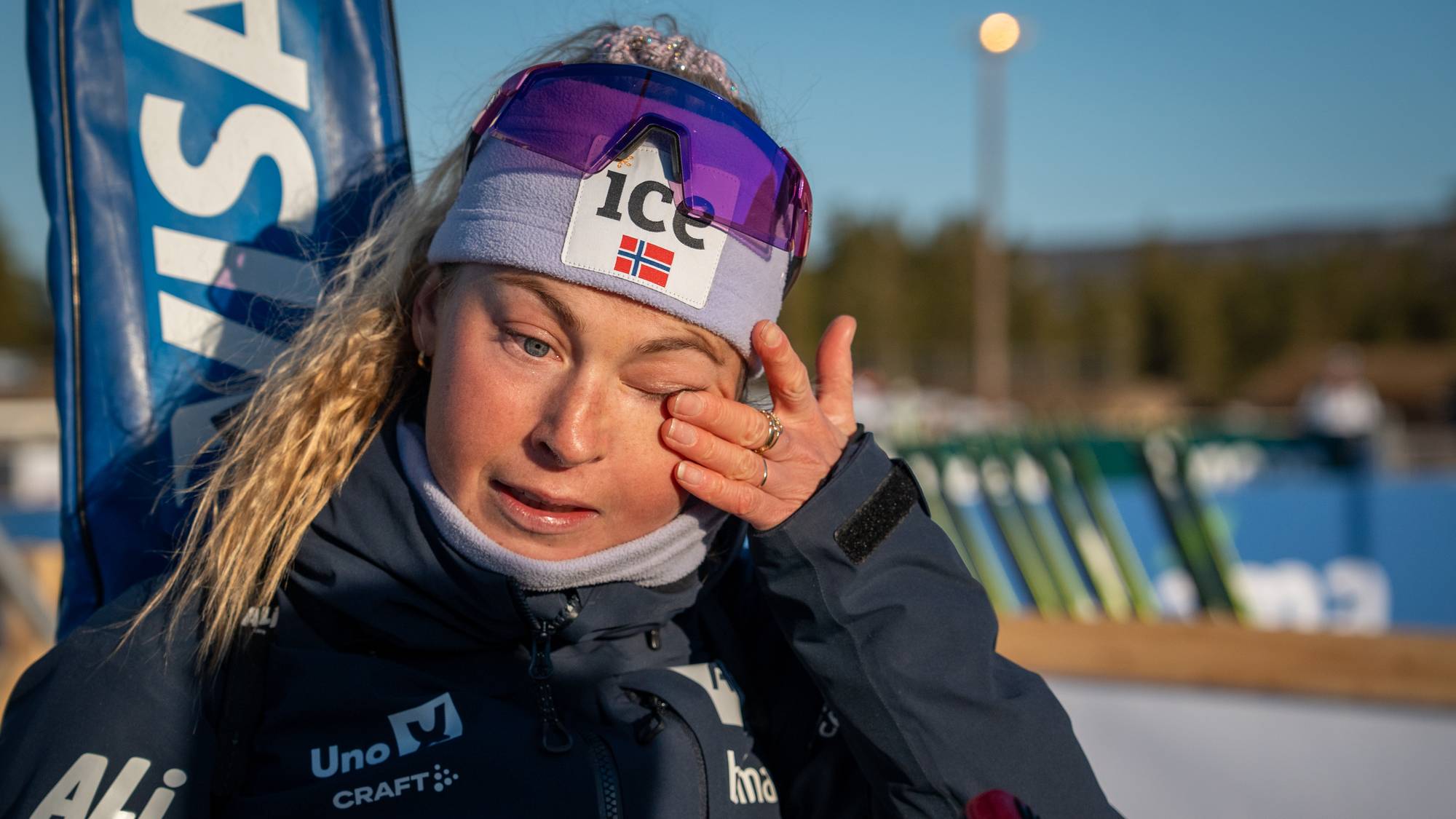 En skiskytter tørker seg i øyet med hånden. Hun er kledd i en blå jakke med sponsorlogoer. I bakgrunnen står ski på stativ. Solen skinner på et snødekt landskap. (Bildebeskrivelsen er laget av en KI-tjeneste)
