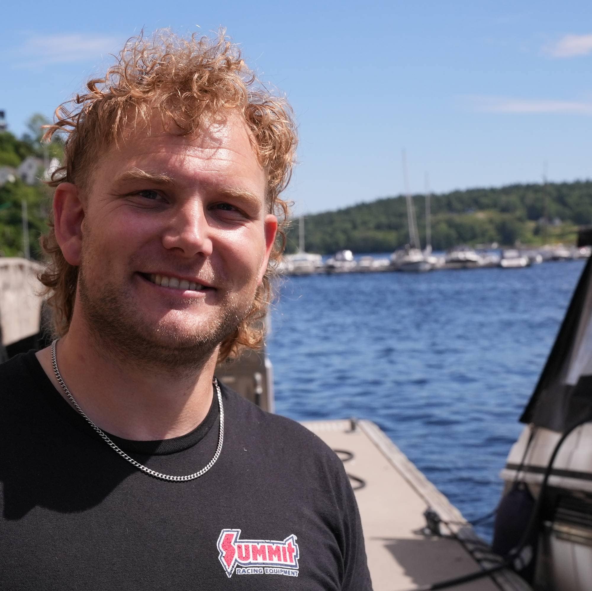 Mann med langt, krølla hår står på brygge med båt og sjø i bakgrunnen. 