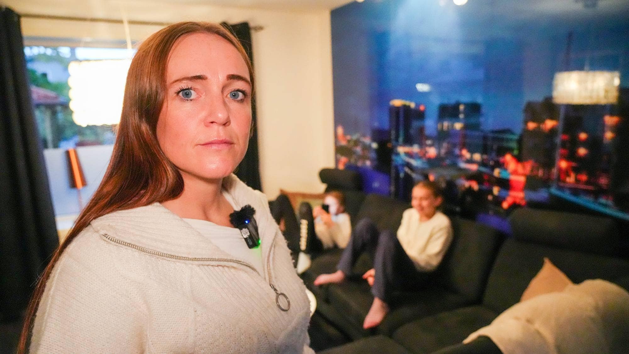 En dame med langt brunt hår og hvit genser ser mot kamera i en stue. I sofaen i bakgrunnen sitter ei jente.