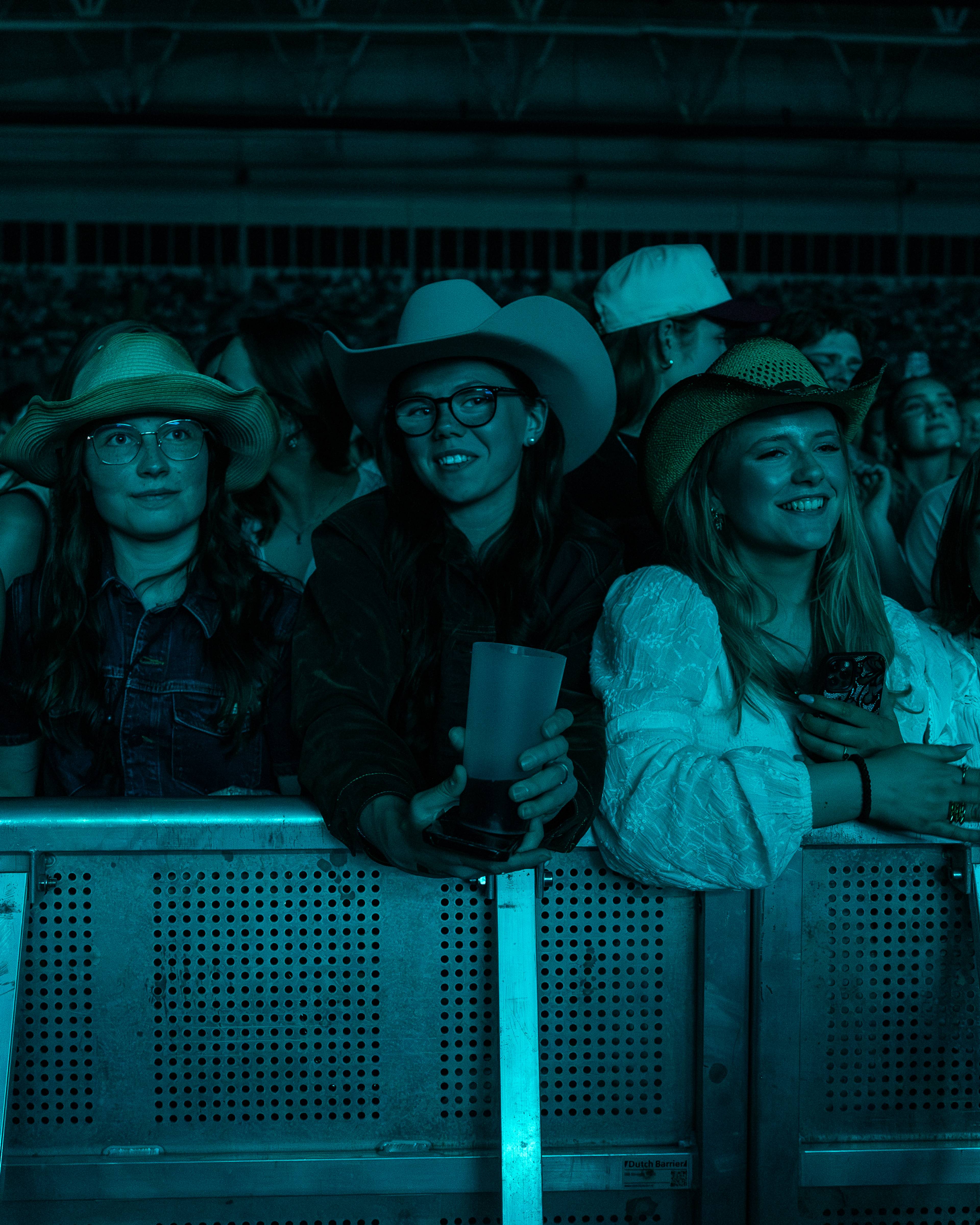 En gruppe unge mennesker står ved et rekkverk under en konsert. De har på seg cowboyhatter og ser ut mot scenen. Bildet er tatt i blått lys. (Bildebeskrivelsen er laget av en KI-tjeneste)