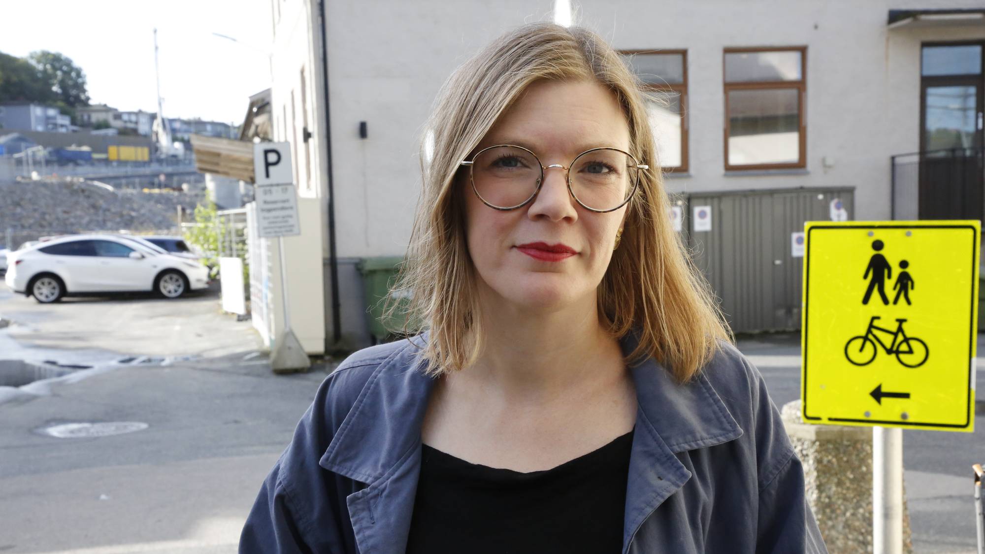Kvinne med briller står utendørs ved et skilt for gående og syklende. (Bildebeskrivelsen er laget av en KI-tjeneste)