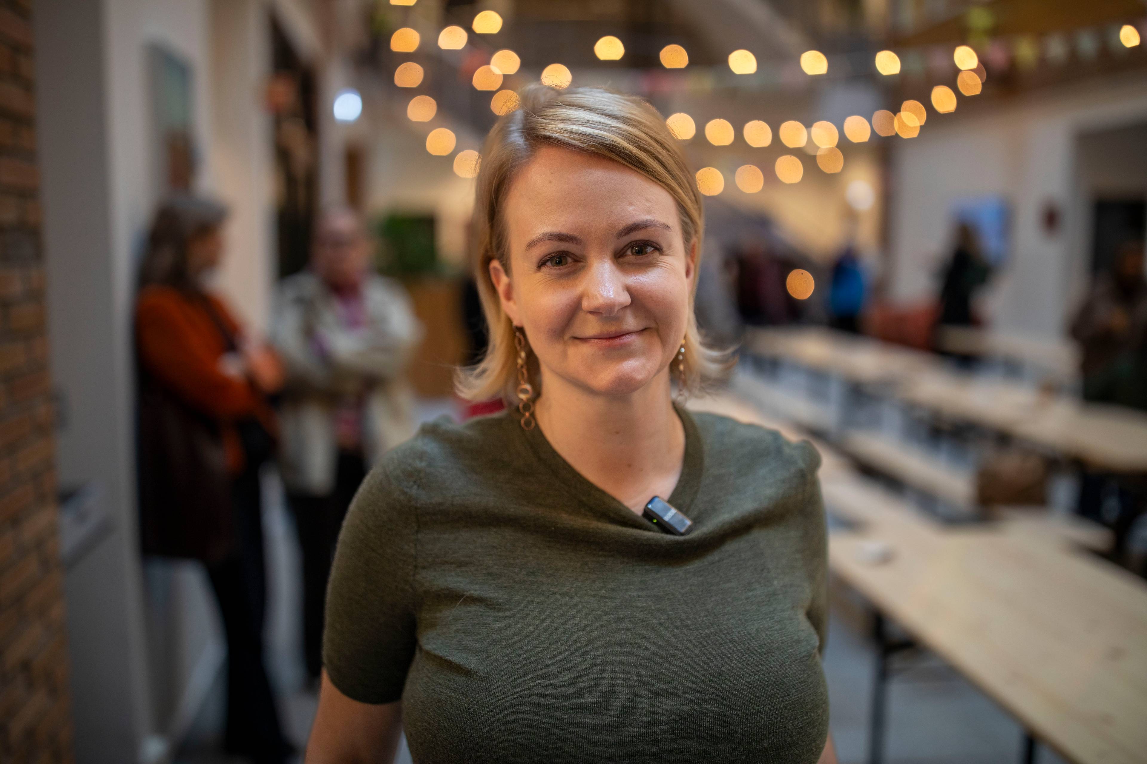Julianne Ferskaug smiler til kameraet. Hun har på seg grønn t-skjorte og har bondt kort hår. Bak henne er det langbord og en del folk i bakgrunnen