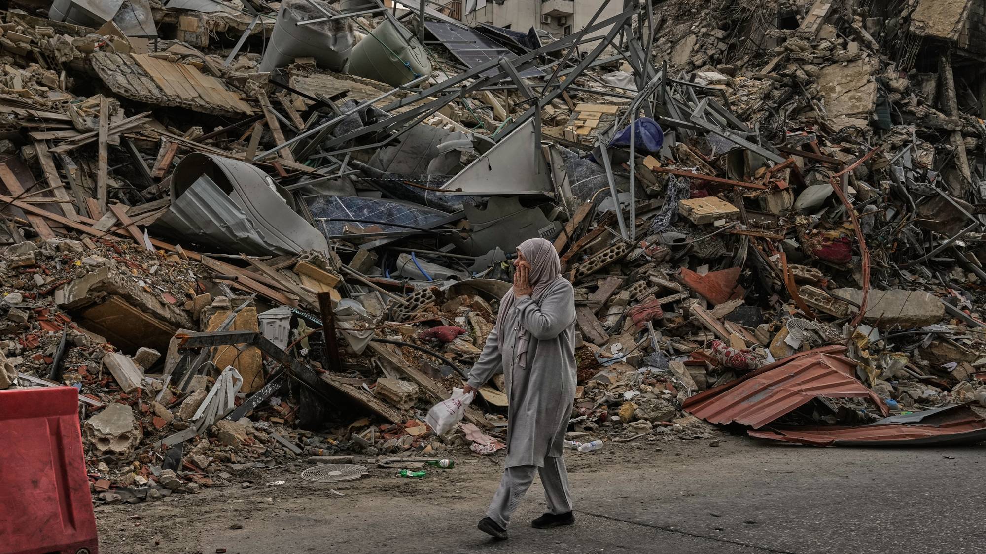 En kvinne med hodeplagg går forbi en stor haug med bygningsrester. Skadene ser ut til å være etter en eksplosjon eller bombe. I bakgrunnen står intakte, men skadede bygninger. (Bildebeskrivelsen er laget av en KI-tjeneste)