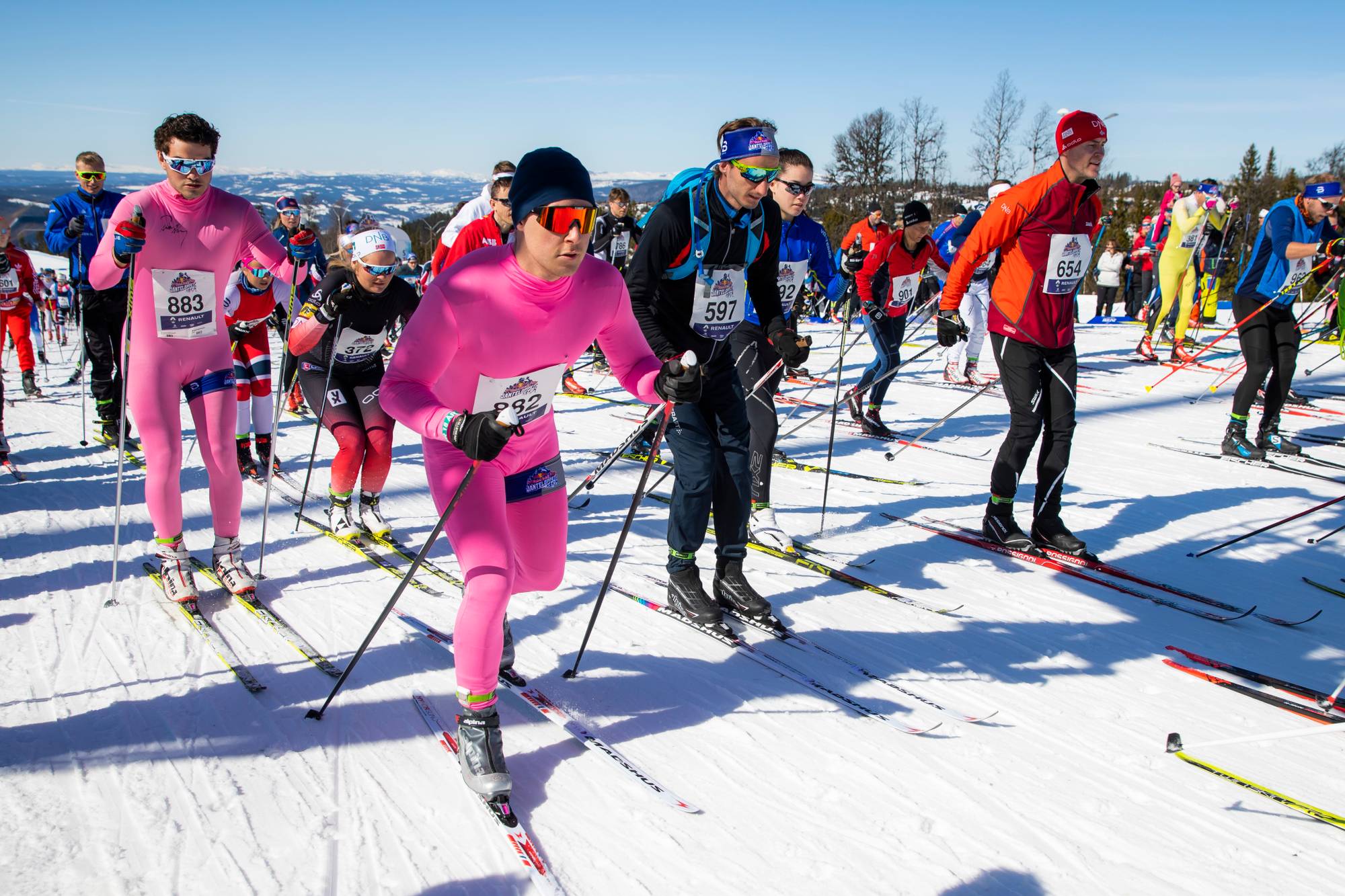 Et stort antall skiløpere står klare ved startstreken i en konkurranse. Flere av deltakerne er iført fargerike dresser, inkludert en gruppe i knallrosa. Solen skinner og det er snødekte fjell i bakgrunnen.