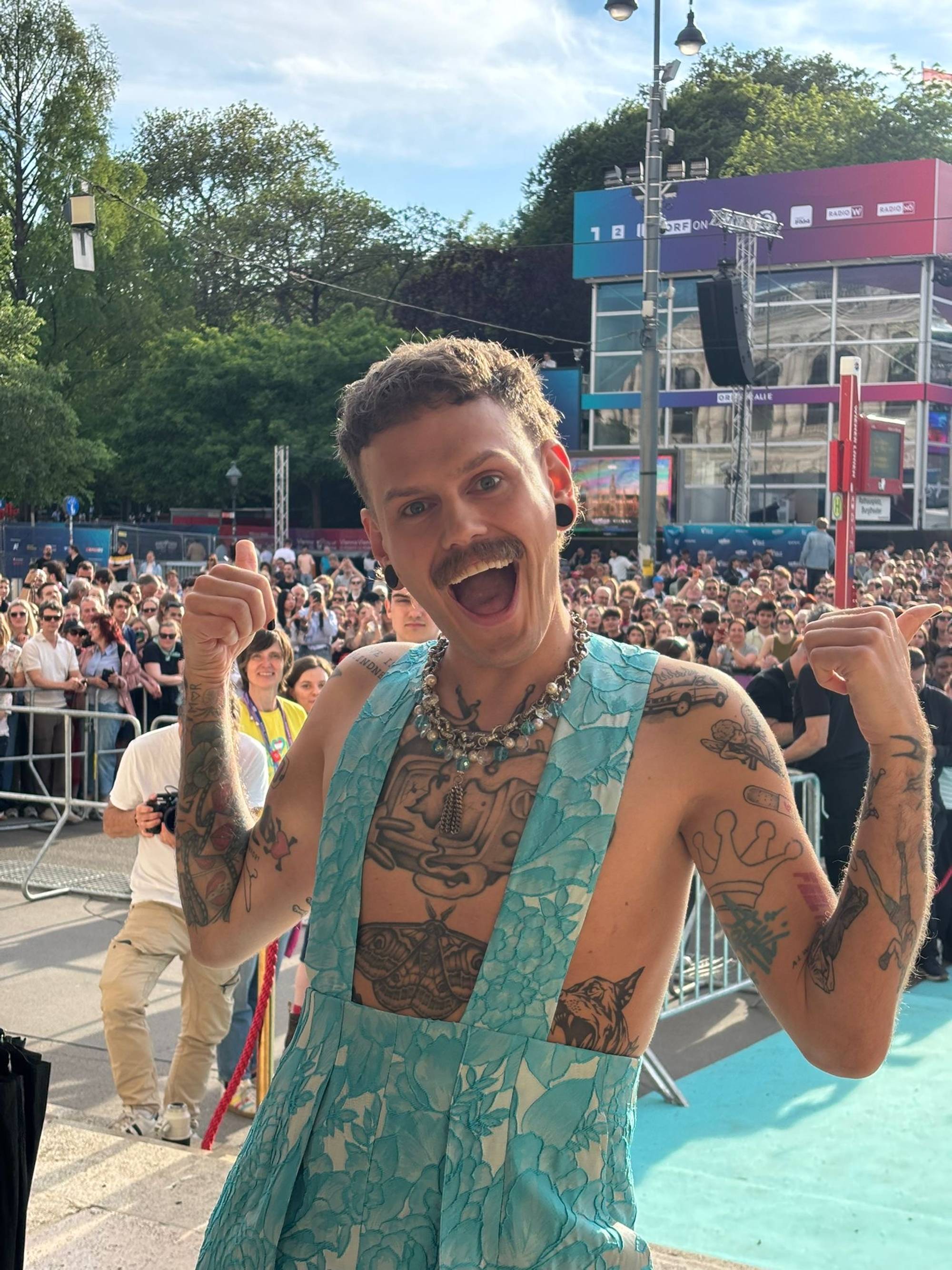 En mann med tatoveringer og en blomstrete jumpsuit gir tommel opp. Han står foran en folkemengde og en scene med et stort fargerikt banner. Mannen har et bredt smil og et smykke rundt halsen. (Bildebeskrivelsen er laget av en KI-tjeneste)