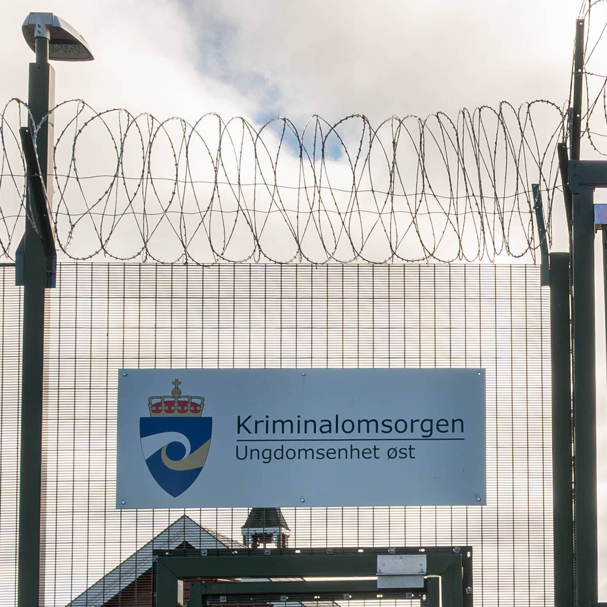 Skilt med Kriminalomsorgen Ungdomsenhet øst montert på et høyt gjerde med piggtråd. (Bildebeskrivelsen er laget av en KI-tjeneste)