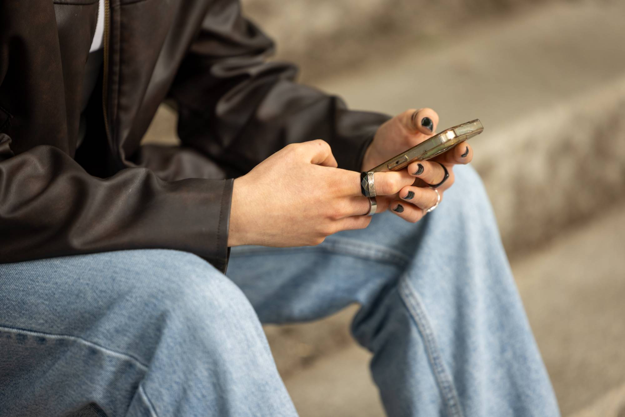 Ung kvinne i skinnjakke og olabukse bruker en mobiltelefon. Hunhar svarte negler og ringer. 