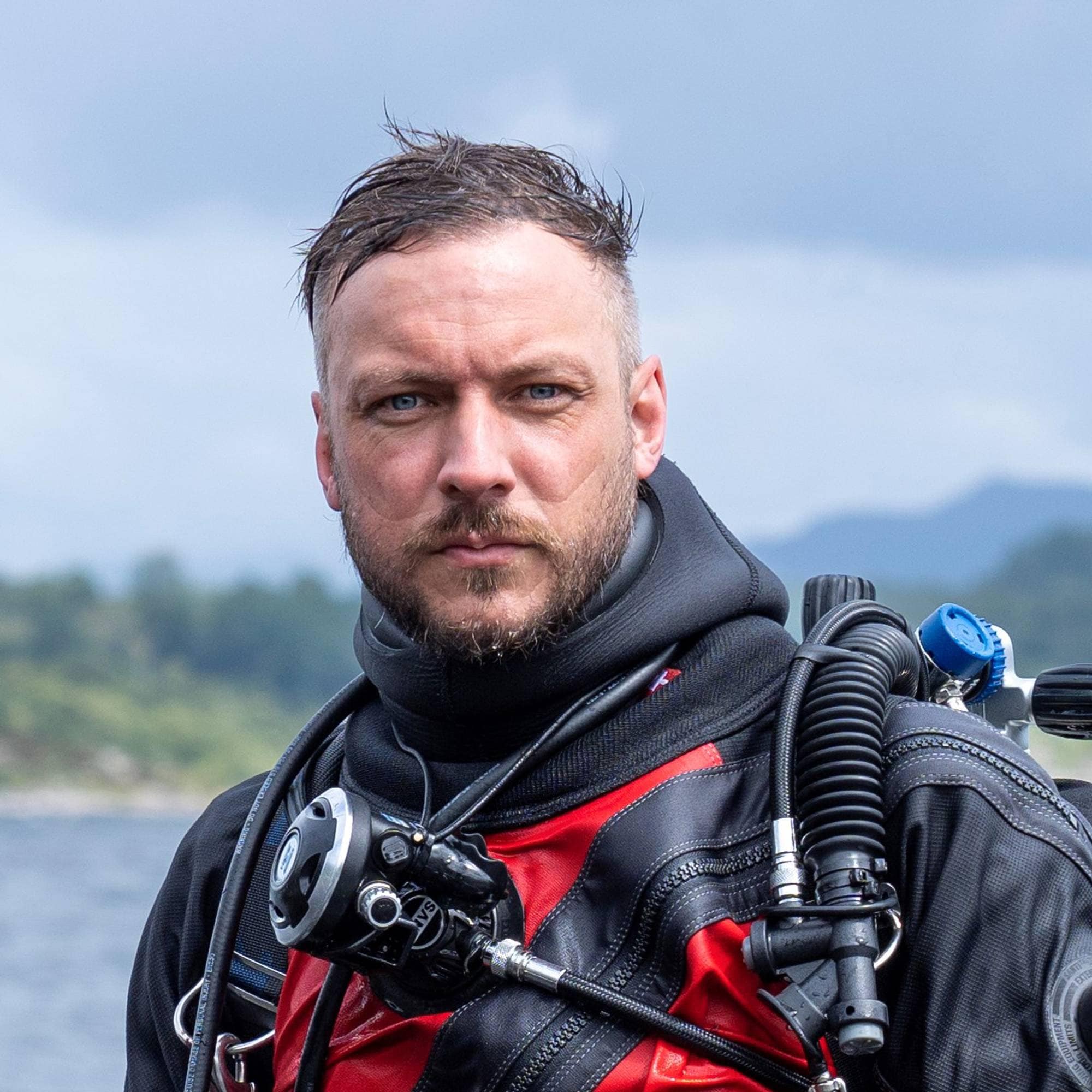 En mann i dykkerutstyr står i vann. Han holder en undervannskamera og et par svømmeføtter. Bakgrunnen er en innsjø med skogkledde åser i det fjerne. (Bildebeskrivelsen er laget av en KI-tjeneste)