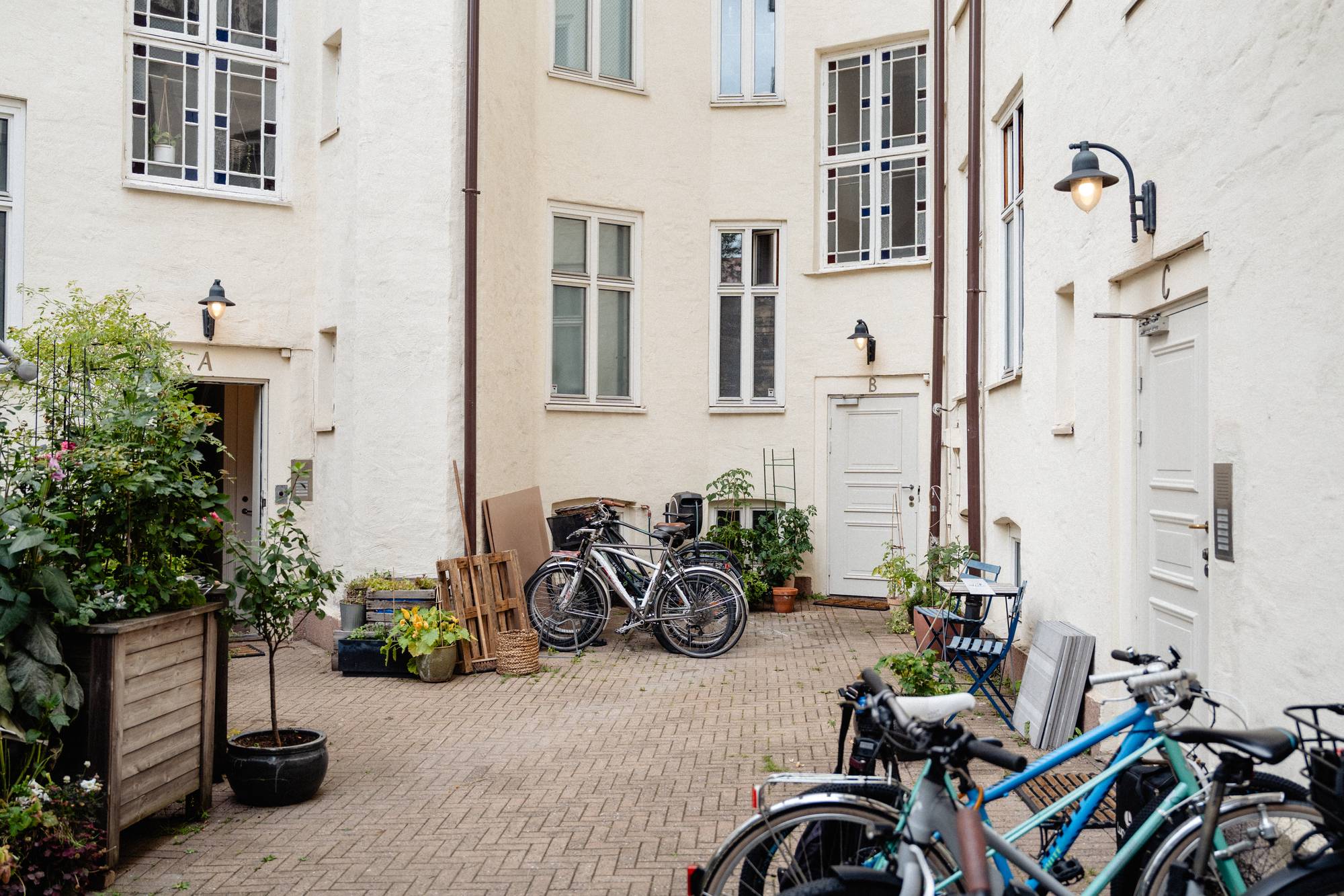 Et hjørne av en bakgård med flere hus satt sammen. Bygården har fine, klassiske vinduer. Sykler er parkert og noen busker står i kasser.