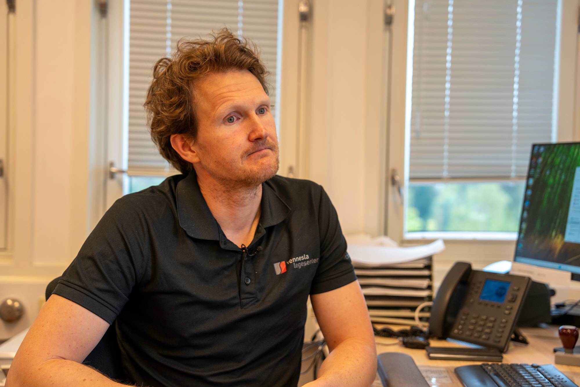 En mann sitter ved et skrivebord på et kontor. Han har på seg en svart pikétrøye med logoen til Vennesla legesenter. Bak ham er det et vindu med persienner. (Bildebeskrivelsen er laget av en KI-tjeneste)