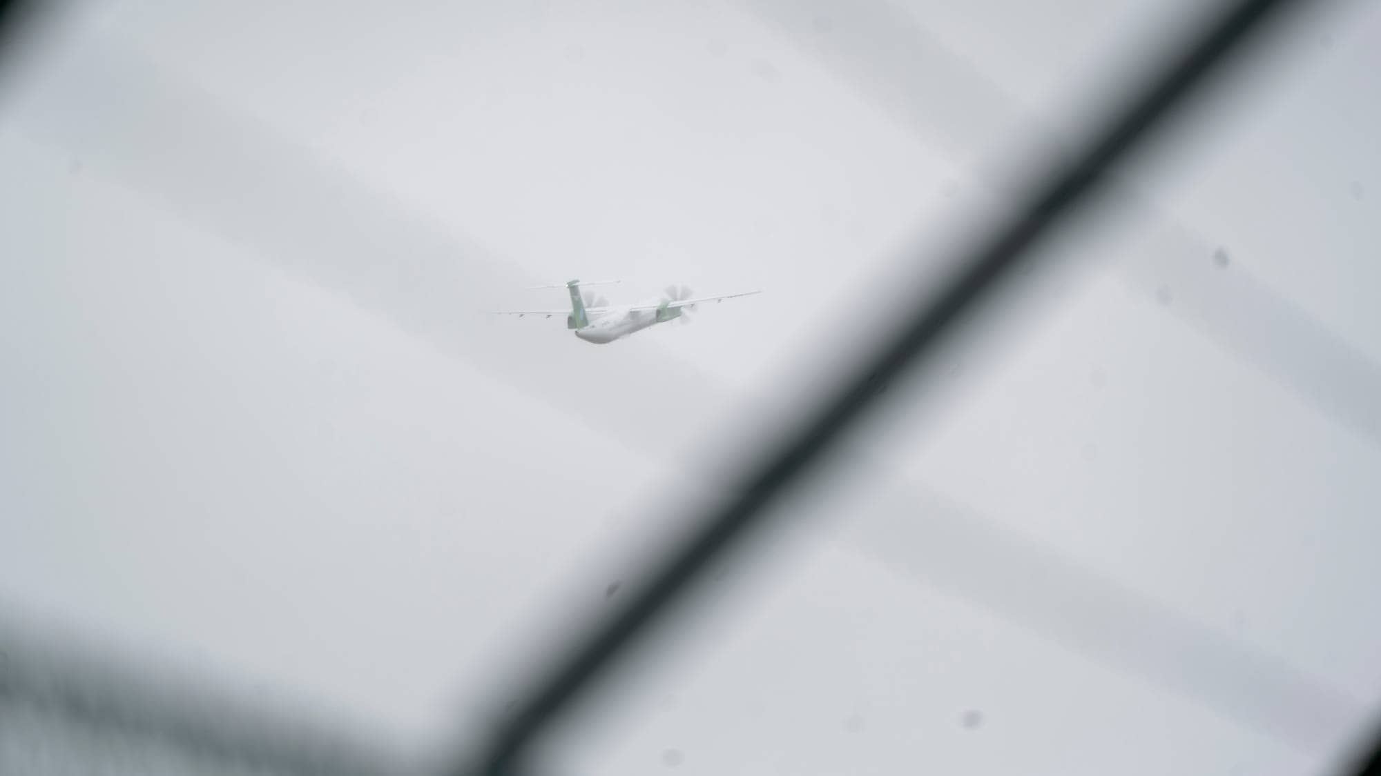 Et propellfly flyr på en grå himmel, sett gjennom et gjerde i forgrunnen. (Bildebeskrivelsen er laget av en KI-tjeneste)