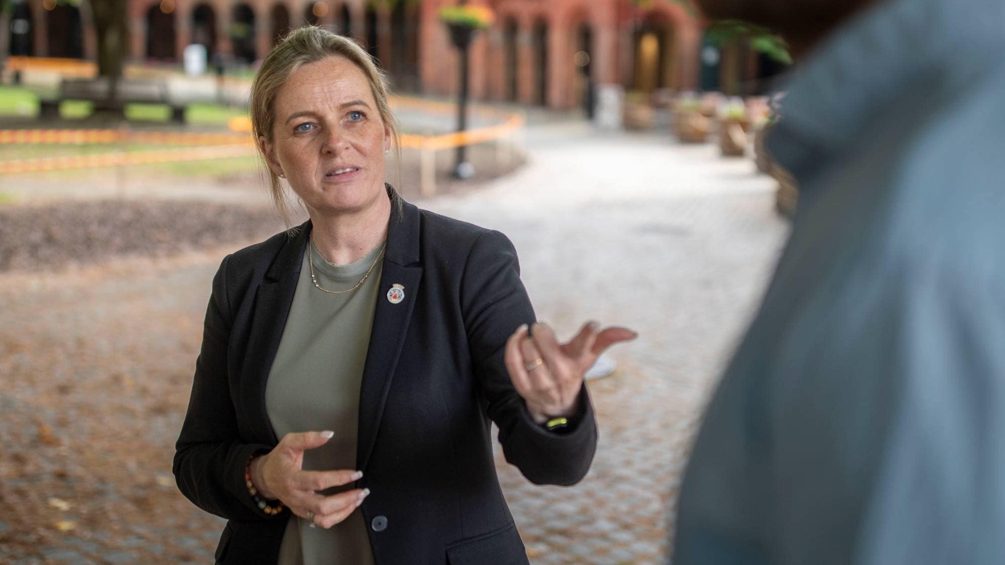 Kvinne i svart jakke snakker og gestikulerer i samtale med en person ut av fokus utendørs. (Bildebeskrivelsen er laget av en KI-tjeneste)