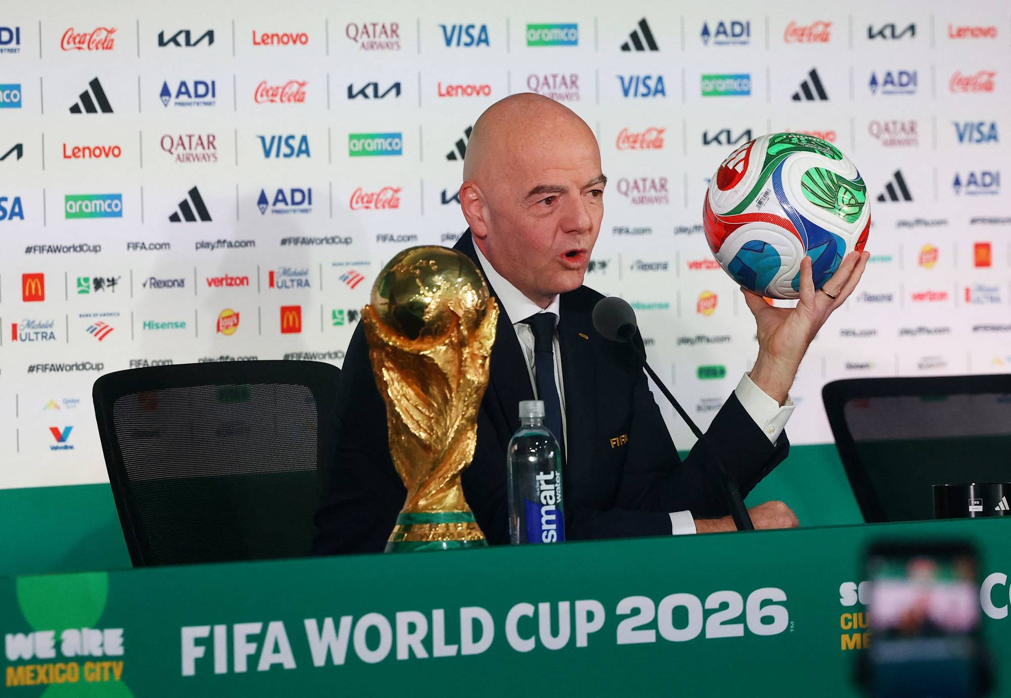 FIFA-president Gianni Infantino holder en fotball og sitter ved siden av VM-trofeet.
Han deltar på en pressekonferanse i forbindelse med VM i fotball.
Bak ham er en bakvegg med flere sponsorlogoer.
En flaske med vann står på bordet foran ham. (Bildebeskrivelsen er laget av en KI-tjeneste)