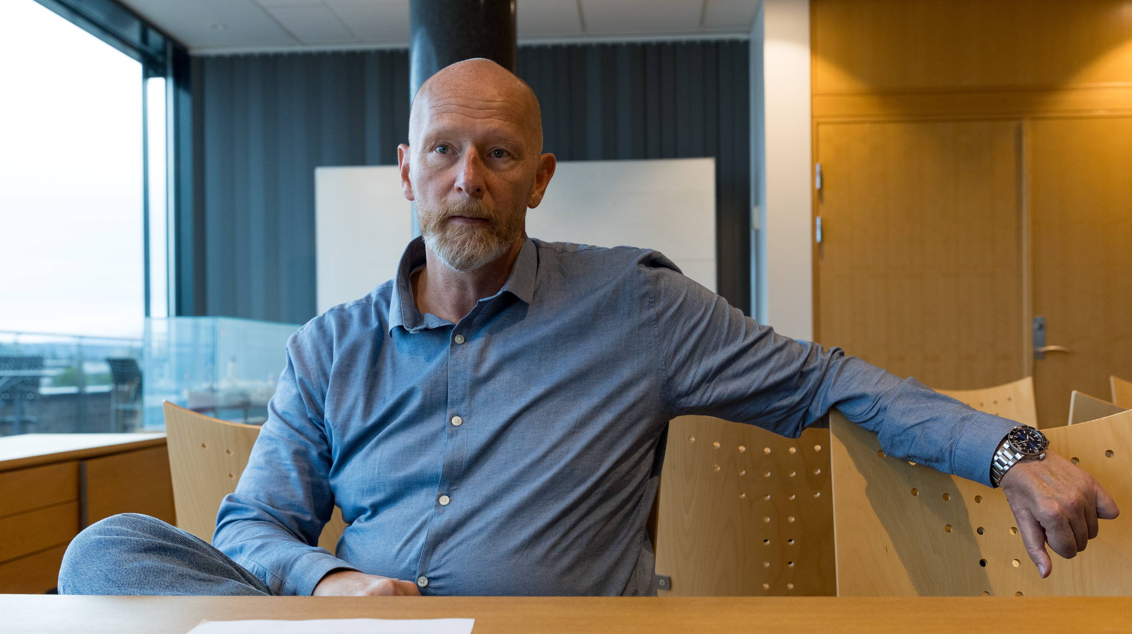 Erik Bjerkaas, svindeljeger i sparebank 1, sitter rundt et bord og lener en arm på stolen ved siden av. Han har skjegg og blå skjorte på. En kan skimte en terasse utenfor vinduet på hans venstre side.
