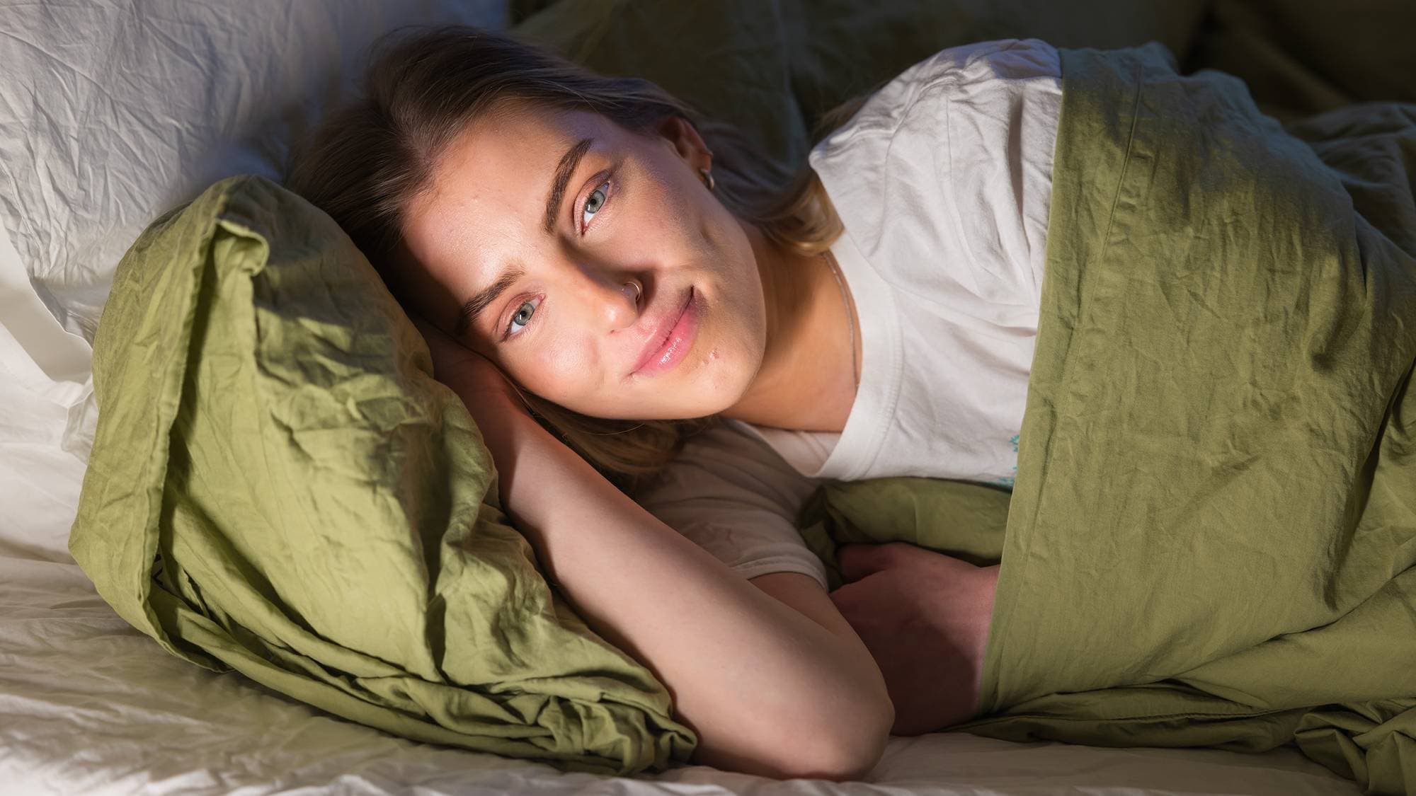 Synne (22) ligger i senga med grønt sengetøy. Hun har høyre arm under hodet, og ser smiler mykt mens hun ser inn i kamera. 
