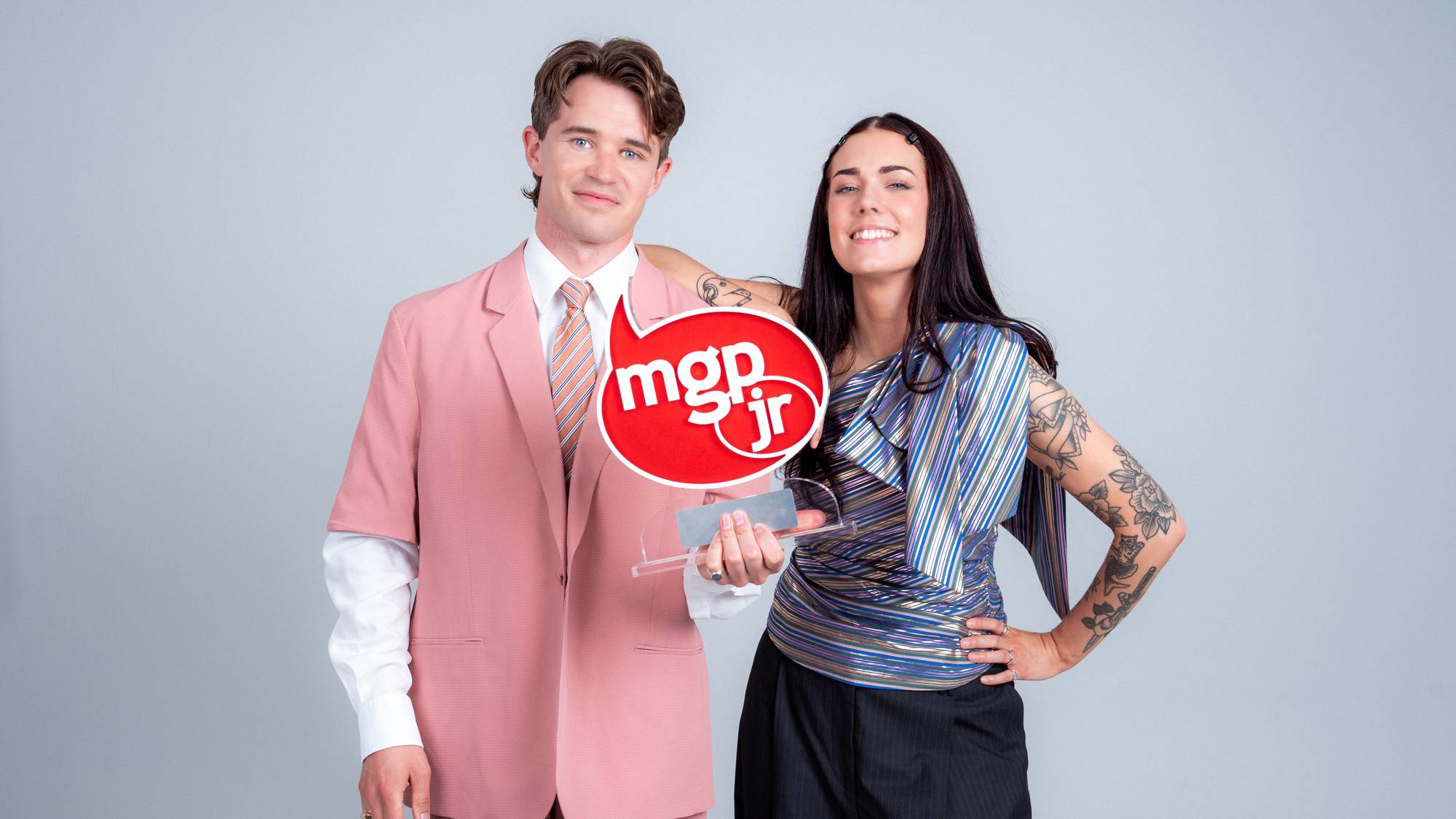 Martin og Tonje holder MGPjr-trofeet. Martin er en mann med brunt hår ikledd rosa dress. Tonje har mørkt, rett hår. Hun har på seg en stripete, glitrende topp og en mørkeblå dressbukse.