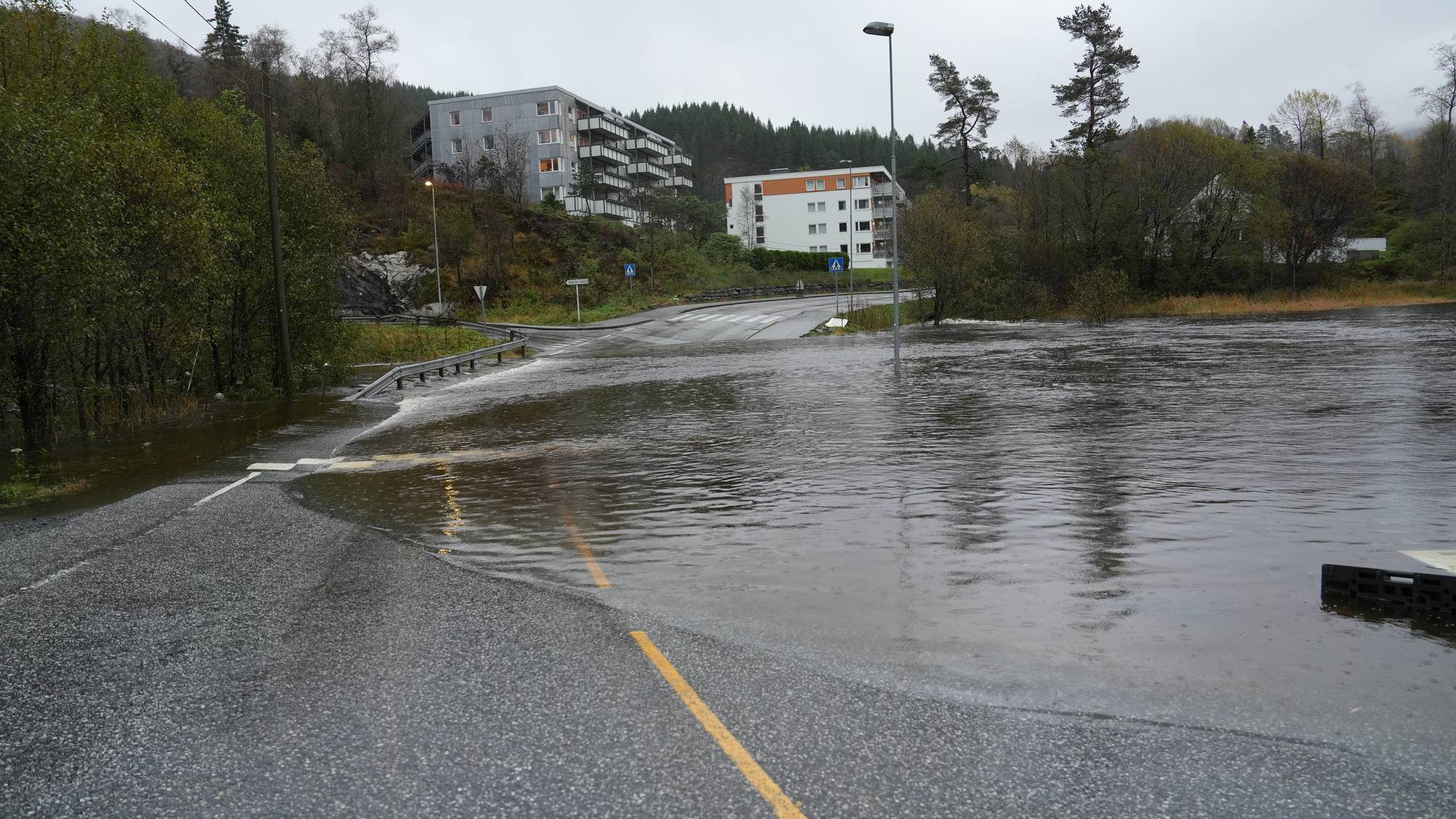 Vannmasser dekker en vei i et boligområde etter flom. Boligblokker ses i bakgrunnen. (Bildebeskrivelsen er laget av en KI-tjeneste)