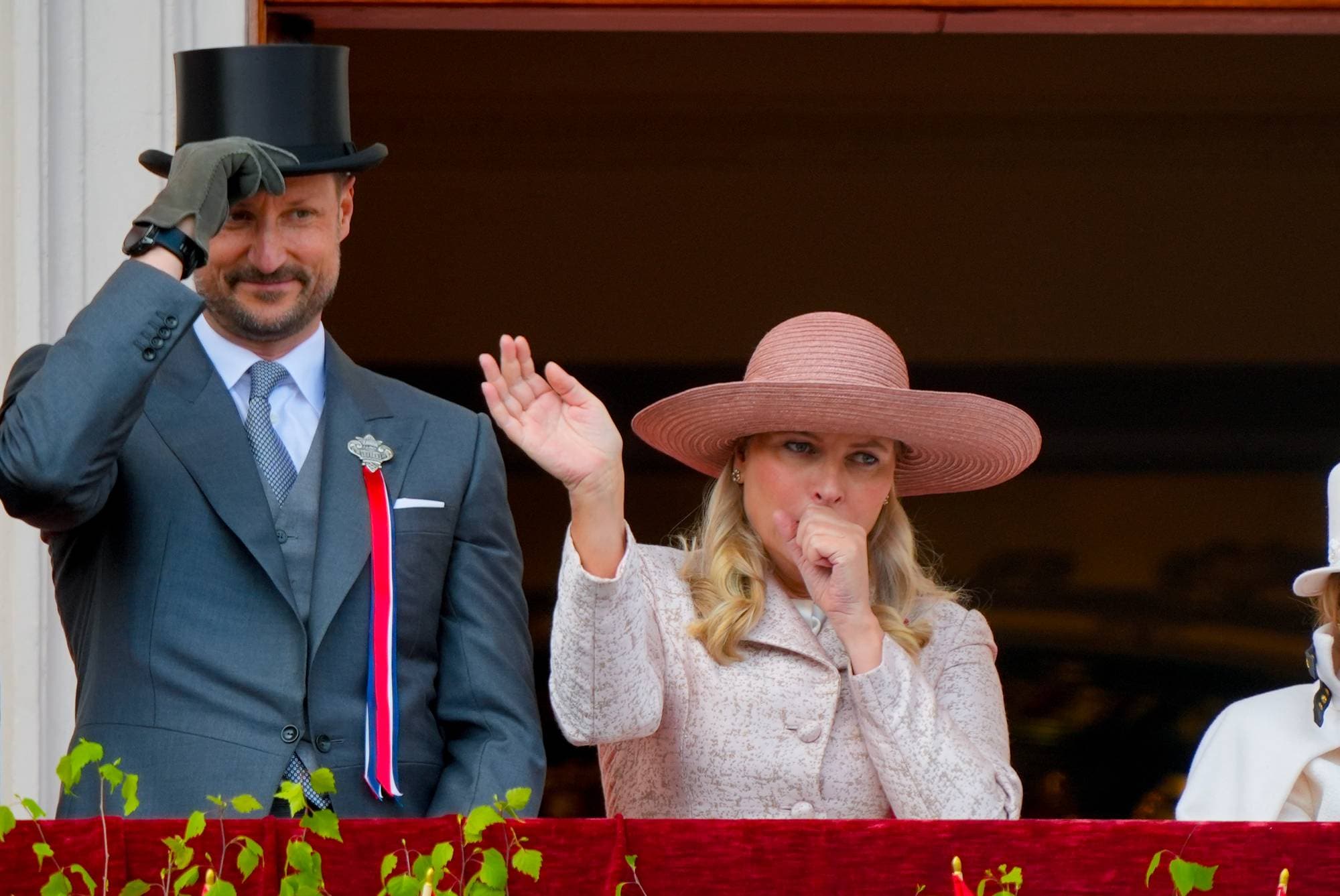 Kronprinsesse Mette-Marit vinker til barnetoget fra Slottsbalkongen mens hun holder seg for munnen og hoster