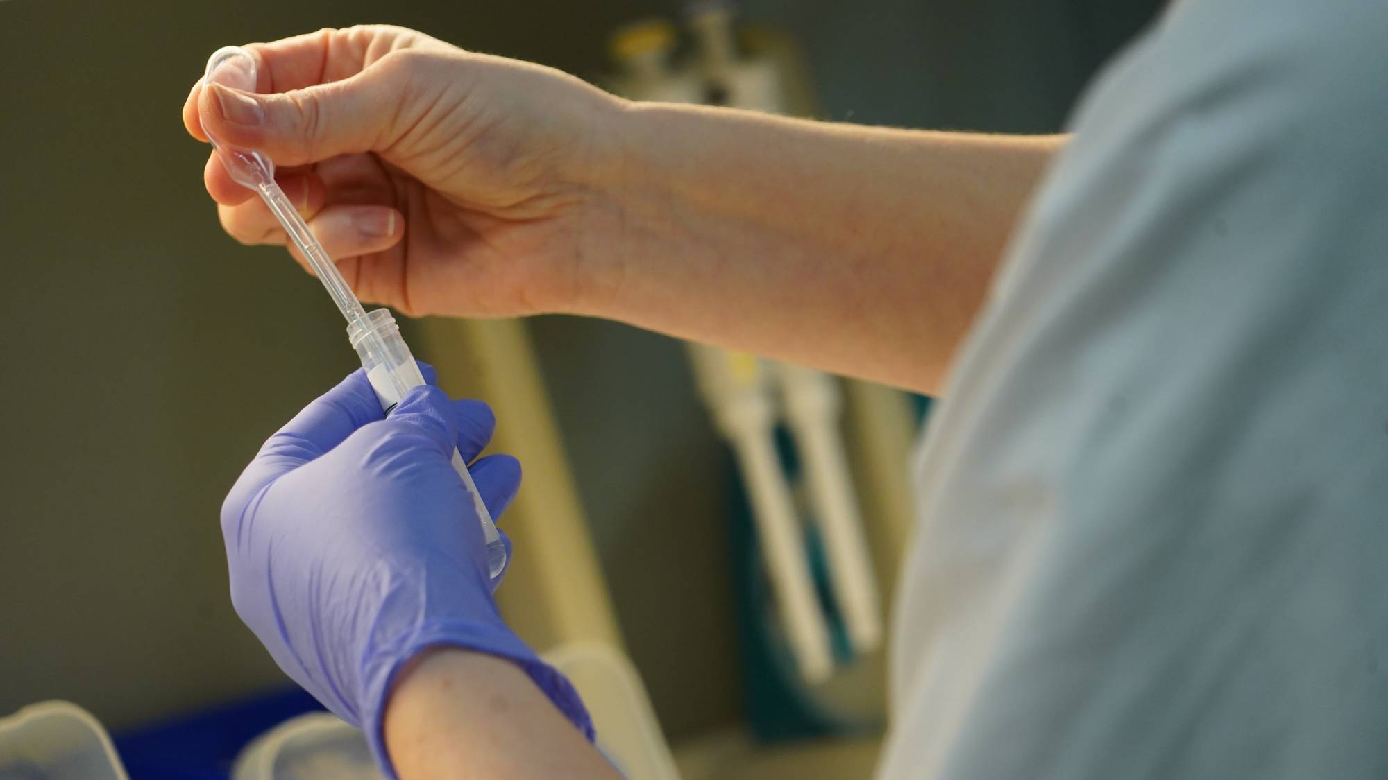 En person iført hansker holder en pipette over et prøveglass. Hendene er fokuspunktet, med tydelig aktivitet knyttet til prøvetaking. Omgivelsene er laboratoriemessige, med pipetter og beholdere synlig i bakgrunnen. Bildet illustrerer et typisk arbeidsmoment i laboratorieforskning. (Bildebeskrivelsen er laget av en KI-tjeneste)
