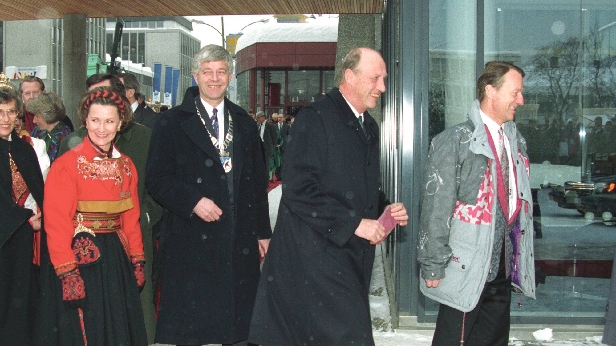 Kong Harald og dronning Sonja på besøk sammen med andre personer på en rød løper utendørs. (Bildebeskrivelsen er laget av en KI-tjeneste)
