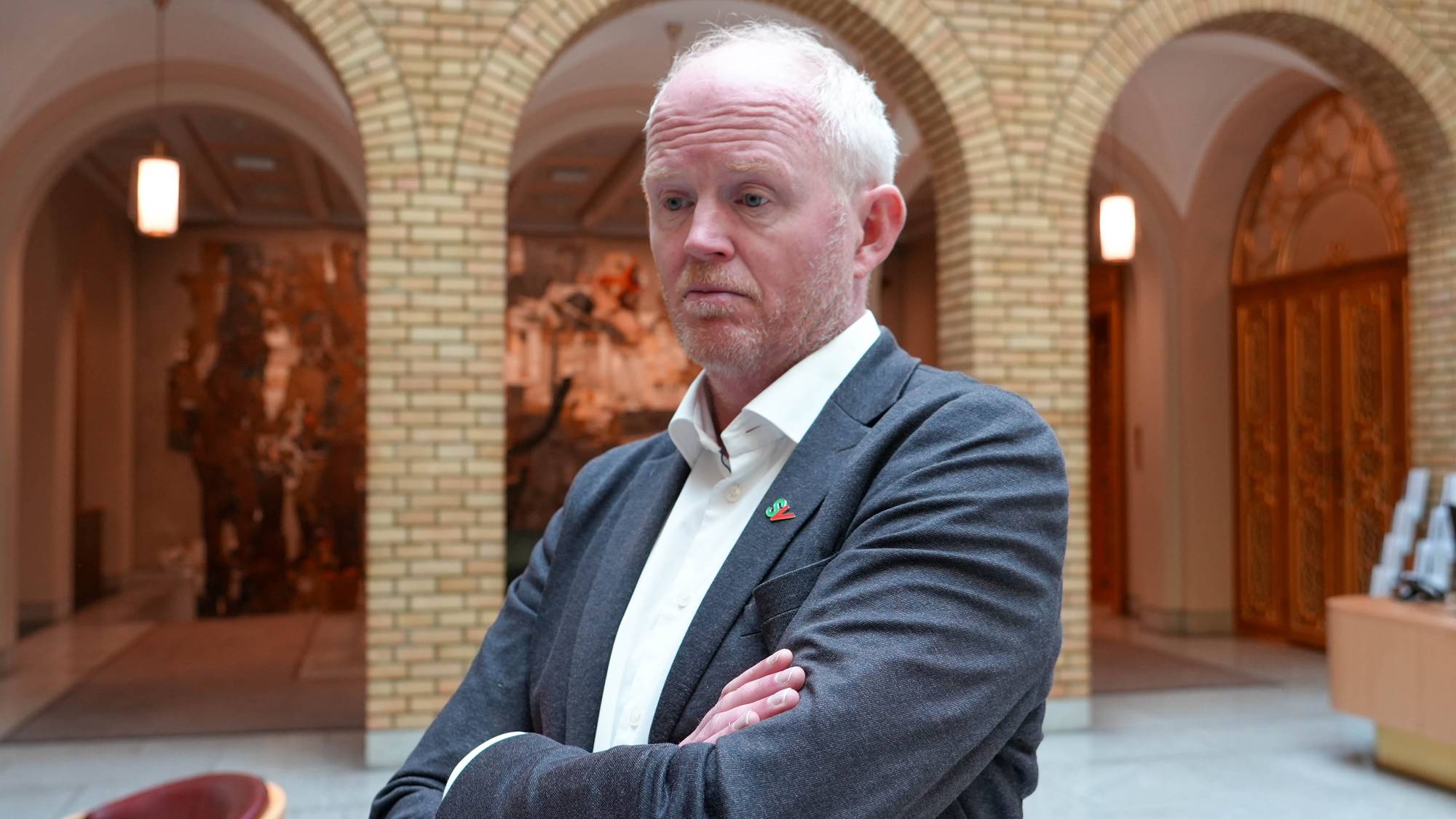 En mann i dress står med armene i kors foran en mursteinsvegg med buer. Han har hvitt hår og et alvorlig uttrykk. (Bildebeskrivelsen er laget av en KI-tjeneste)