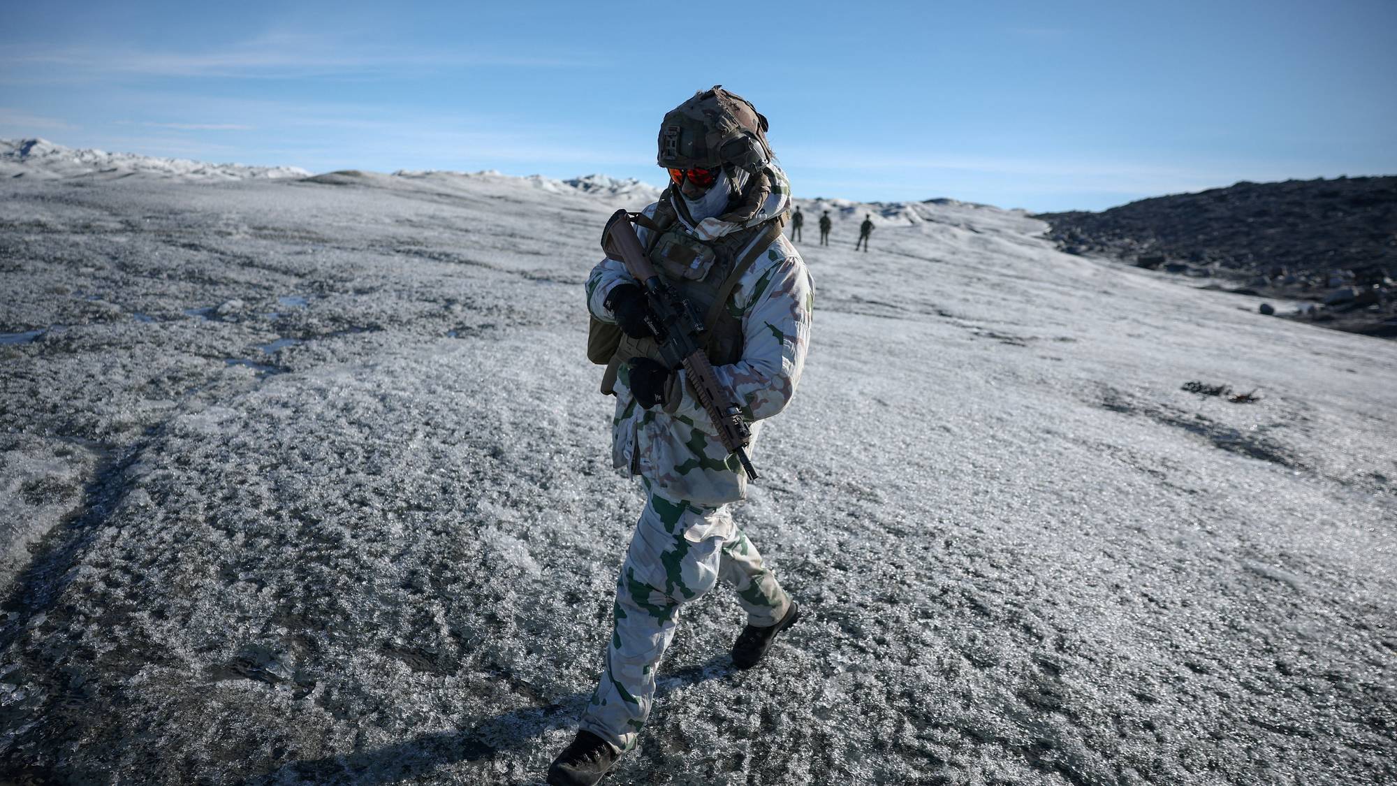 Soldat i kamuflasjeutstyr går med våpen over et snødekt landskap. Flere soldater sees i bakgrunnen. 