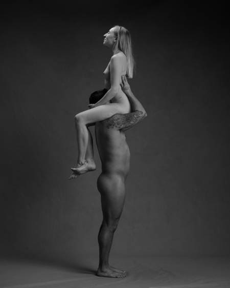 En mann med mørkt hår og tatoveringer står oppreist og bærer en blond kvinne på skuldrene. Hun sitter med skrittet mot ansiktet hans. Begge er nakne, men ingen kjønnsorganer er synlige.