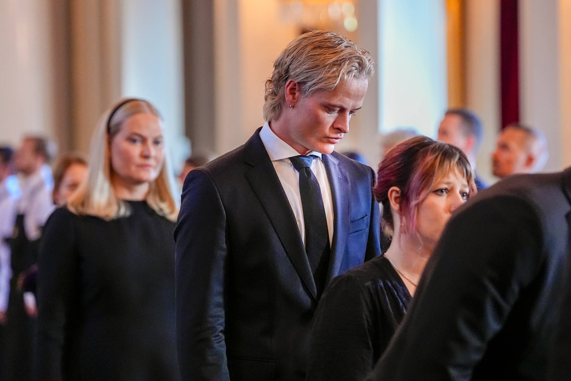 Marius Borg Høiby og kronprinsesse Mette-Marit i et formelt rom med flere andre personer. 