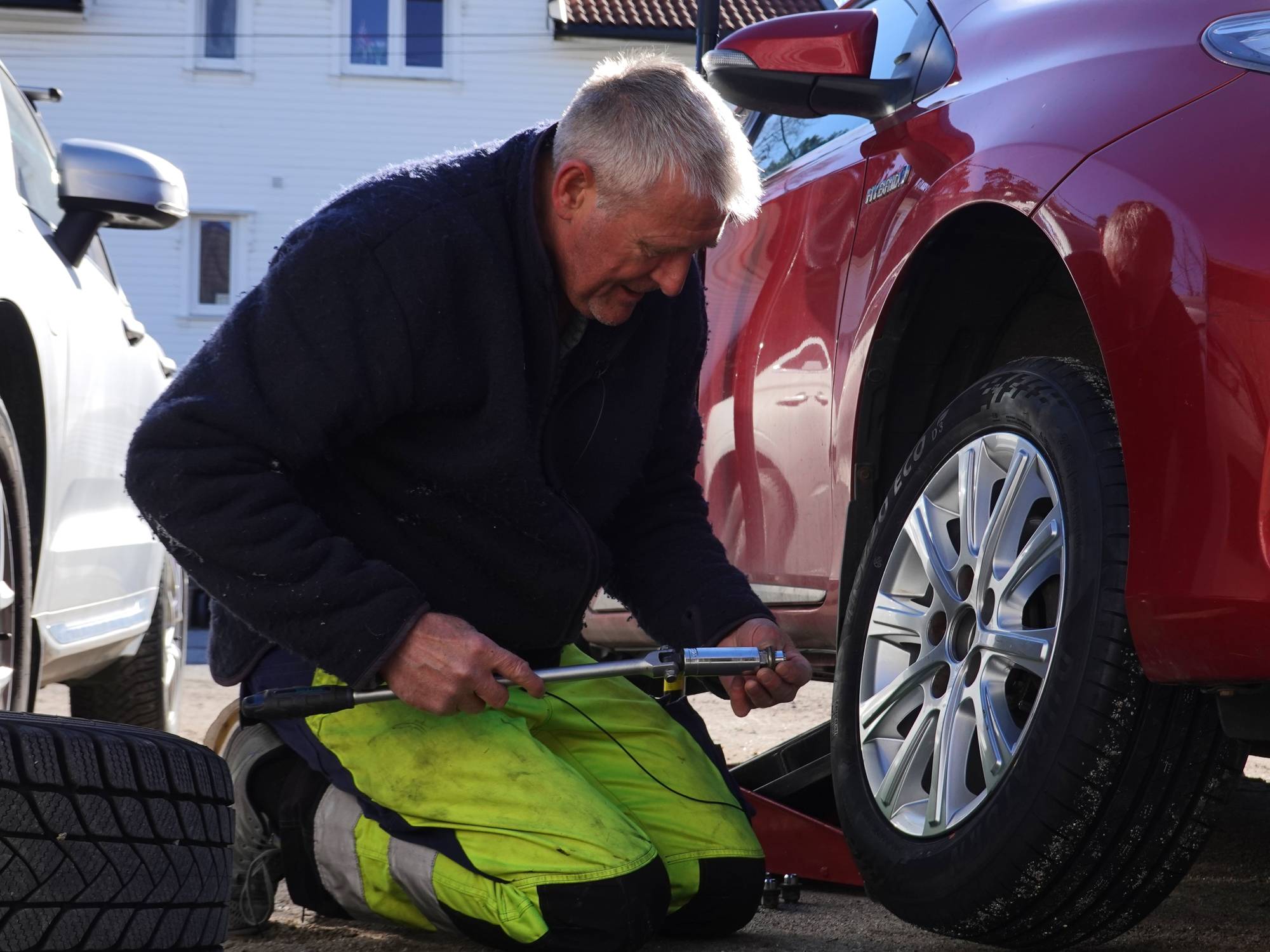 En mann skifter dekk på en rød bil. Han bruker en jekk og et verktøy for å løsne boltene. I bakgrunnen står en annen bil og et hvitt hus. Det er solskinn og klarvær. (Bildebeskrivelsen er laget av en KI-tjeneste)