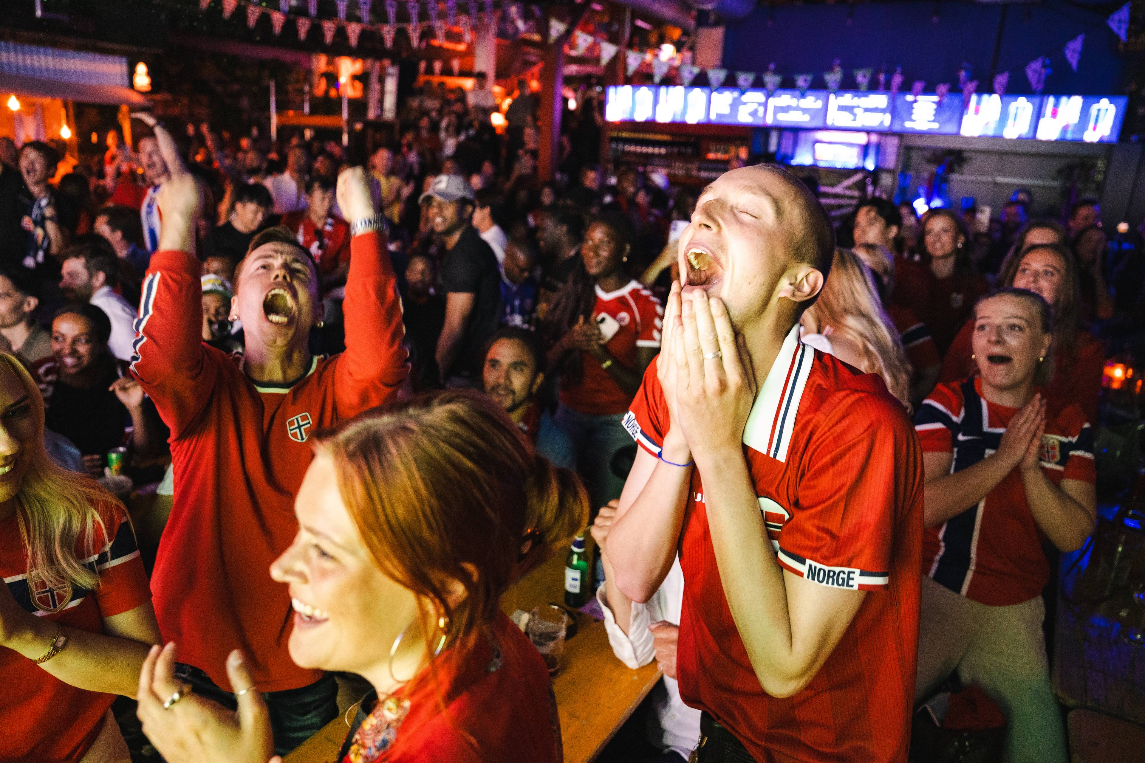 Publikum i norske landslagsdrakter jubler begeistret i en bar under en sportssending. (Bildebeskrivelsen er laget av en KI-tjeneste)
