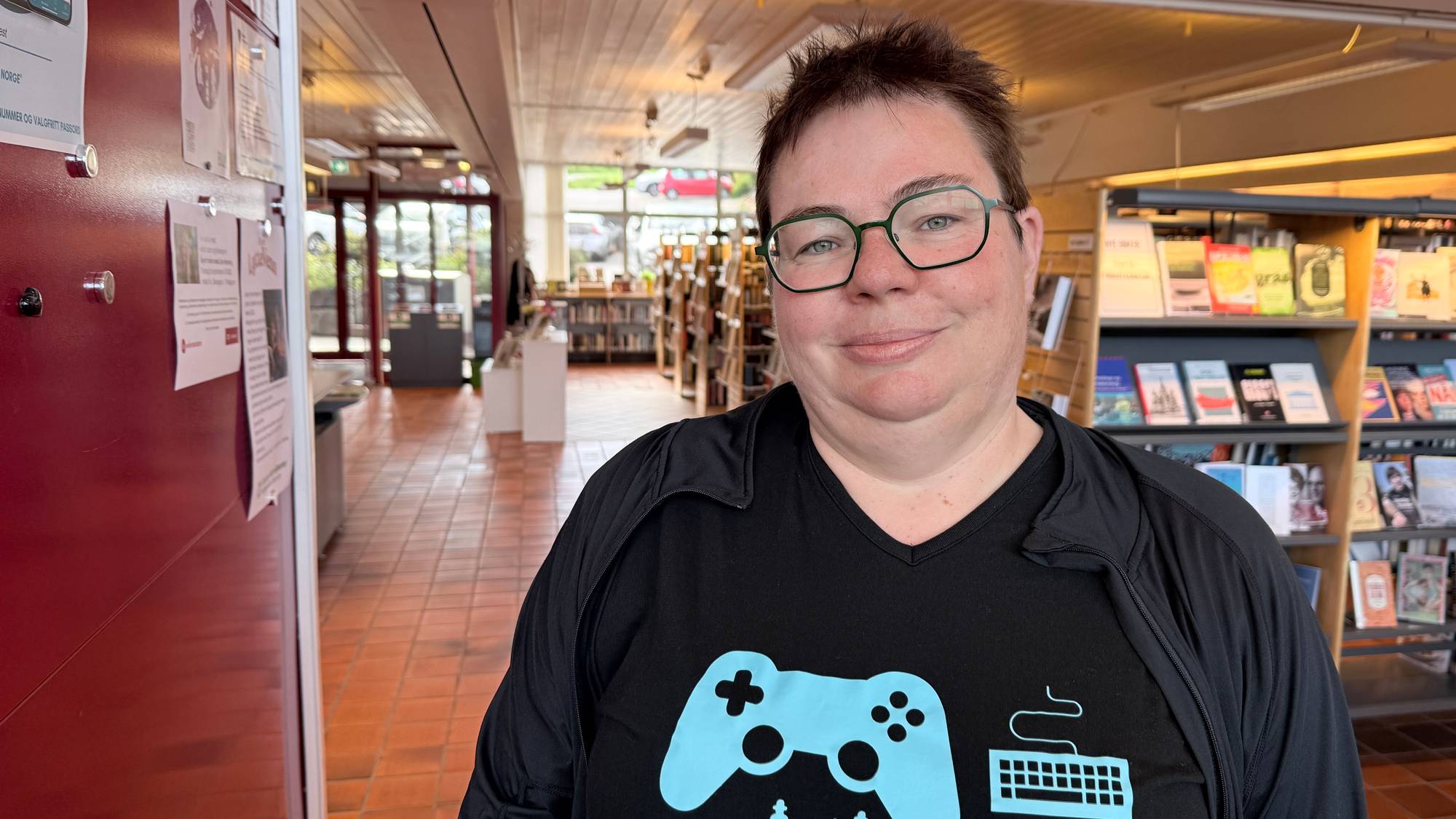 En person med briller og t-skjorte med spillmotiv står inne i et bibliotek. (Bildebeskrivelsen er laget av en KI-tjeneste)