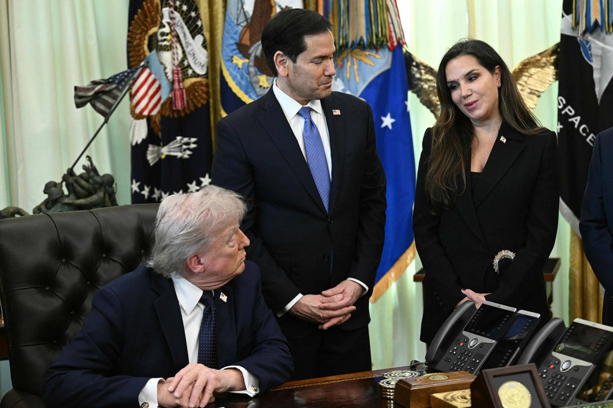 Donald Trump sitter ved et skrivebord med tre andre personer stående bak ham. Blant dem er Marco Rubio. Kvinnen til høyre for Rubio ser ned mens hun snakker. Bak dem er det flere flagg, inkludert et amerikansk flagg og et flagg med en ørn. De er i et kontor som ser ut til å være det ovale kontor. (Bildebeskrivelsen er laget av en KI-tjeneste)