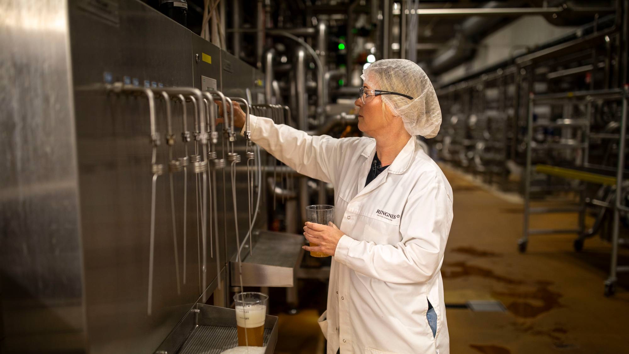 Kvinne i hvit frakk og hårnett tapper øl fra en ølkran inne på en fabrikk. 