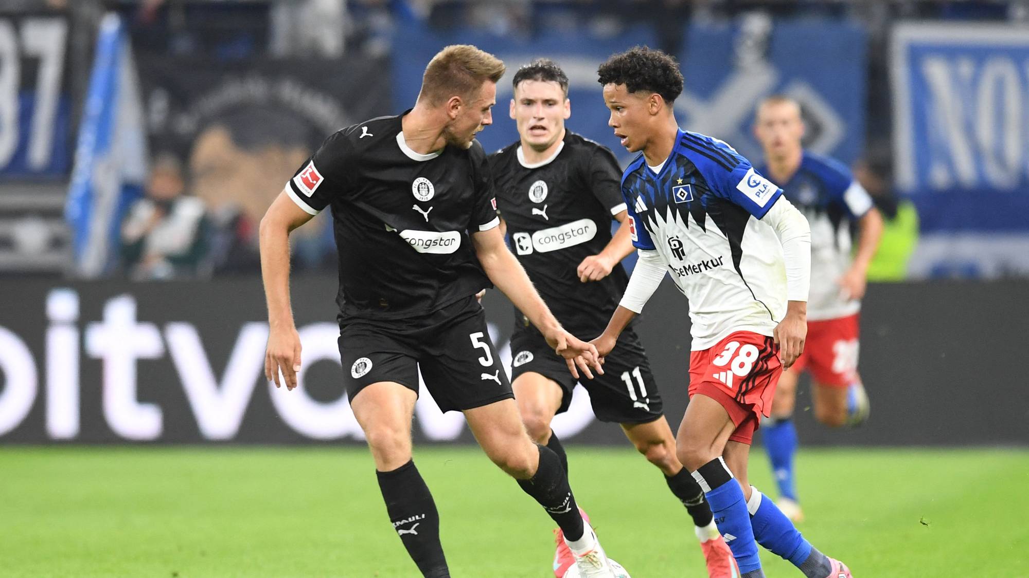 Fotballspillere i aksjon under en kamp. En spiller i svart drakt forsøker å takle ballen fra en spiller i hvit og blå drakt. To andre spillere er i bakgrunnen. Kampen spilles på en gressbane med tilskuere i bakgrunnen. (Bildebeskrivelsen er laget av en KI-tjeneste)