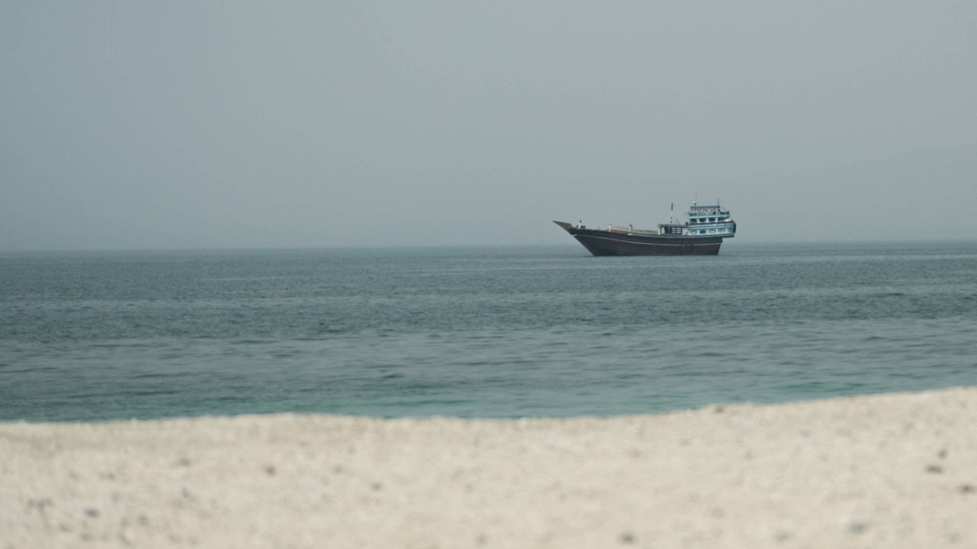 En båt ligger ute på havet, sett fra en strand med lys sand i forgrunnen. (Bildebeskrivelsen er laget av en KI-tjeneste)